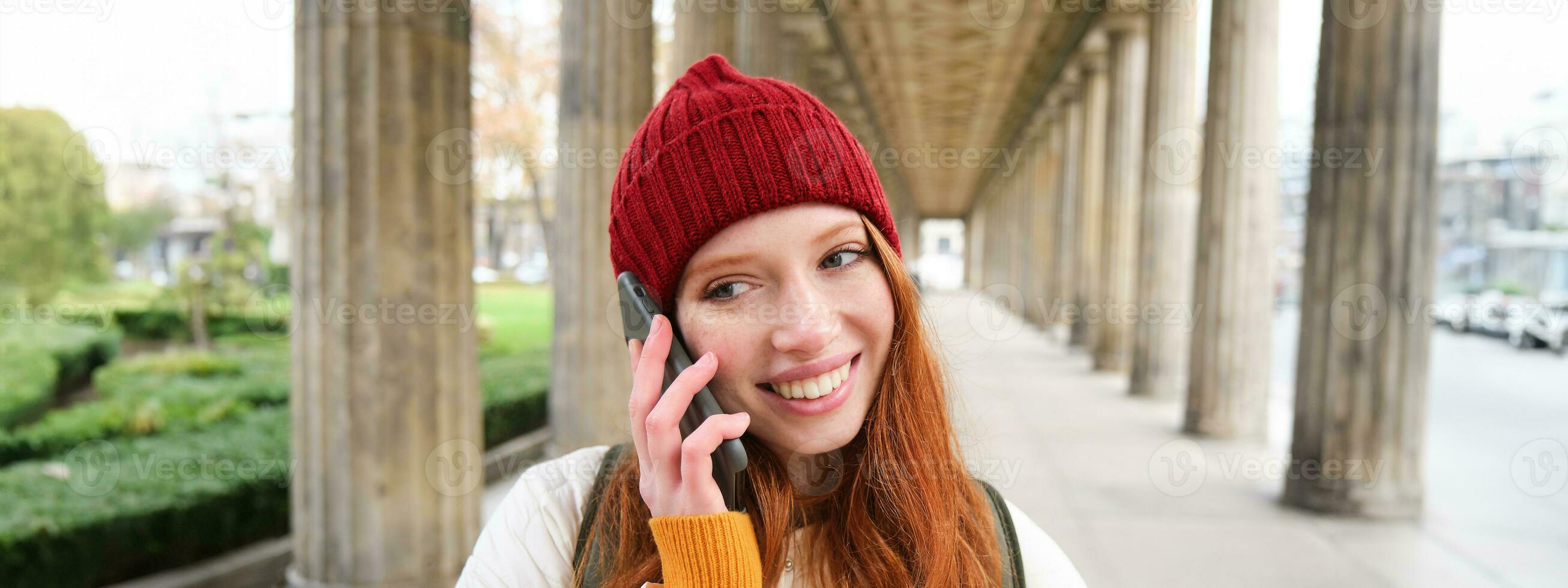 sorridente carino testa Rossa donna fa un' Telefono chiamata, detiene telefono vicino anno, ha mobile conversazione, utilizzando smartphone su strada foto