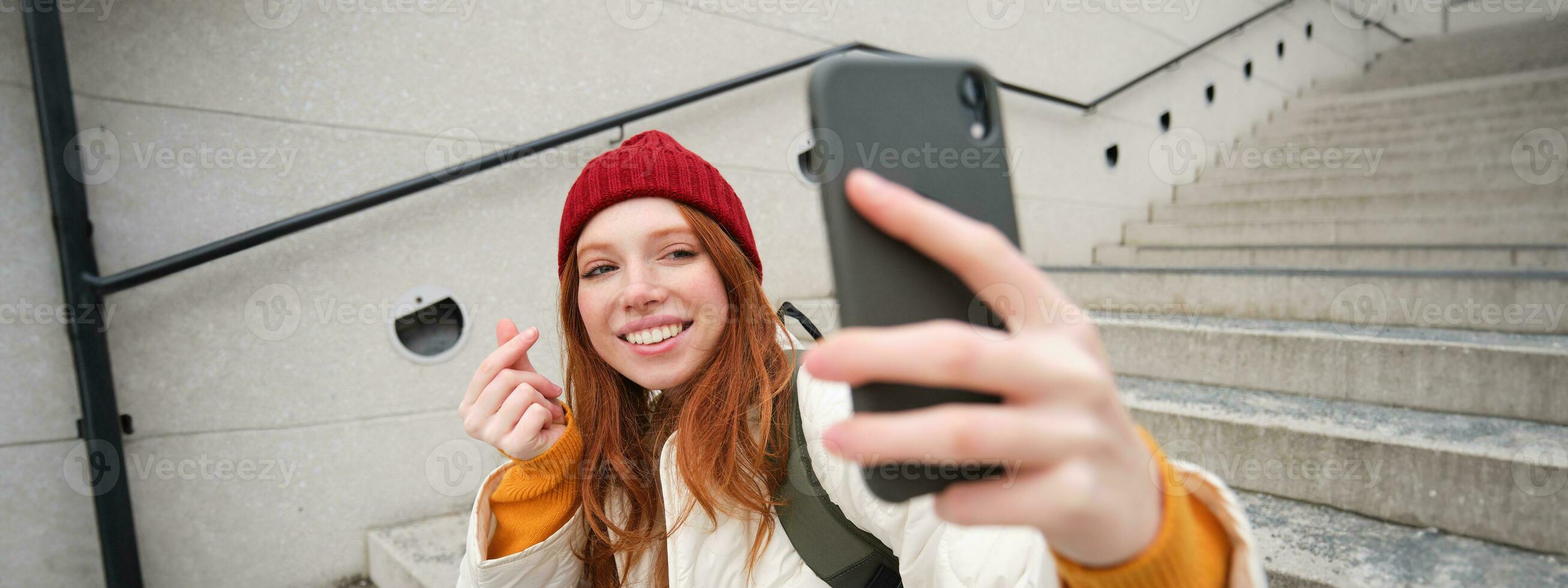 mobile Telefono e persone stile di vita. elegante testa Rossa ragazza prende autoscatto su sua smartphone, pose per foto con mobile Telefono nel mano, sorrisi felicemente, si siede su le scale all'aperto