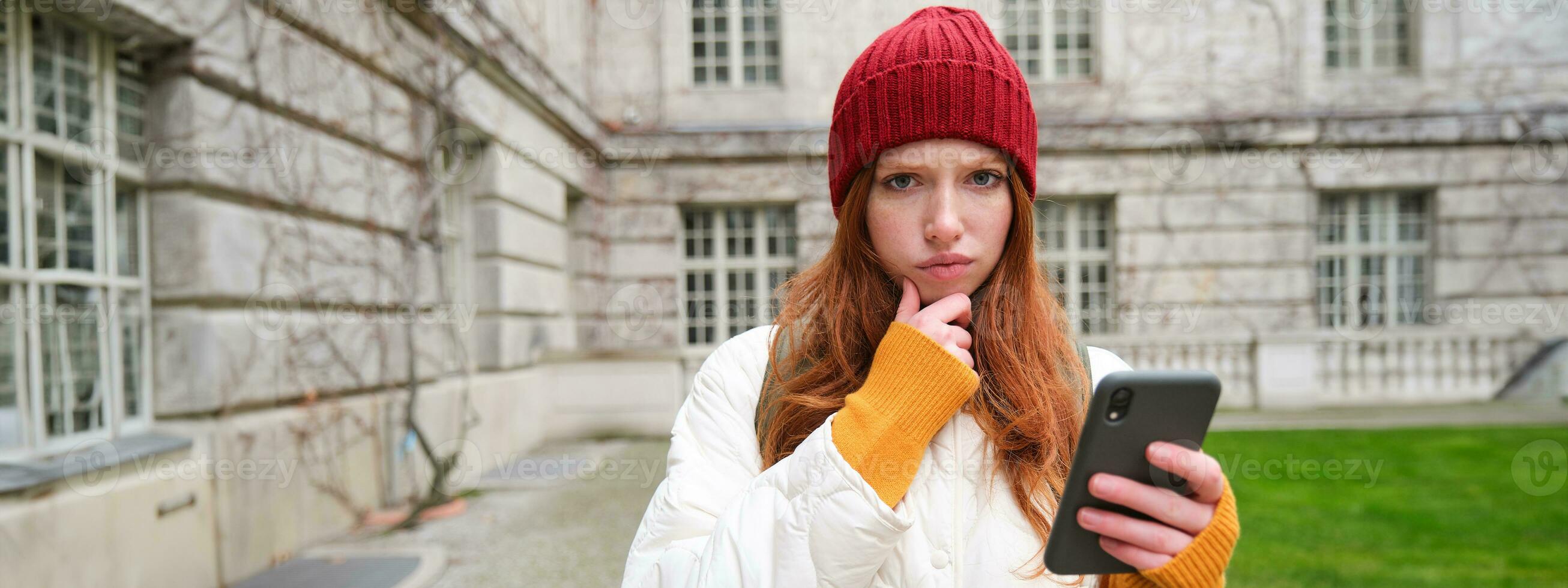 ritratto di giovane testa Rossa donna nel rosso cappello, detiene smartphone e sembra premuroso, accigliato con perplesso, pensiero viso espressione, in piedi su strada vicino edificio foto