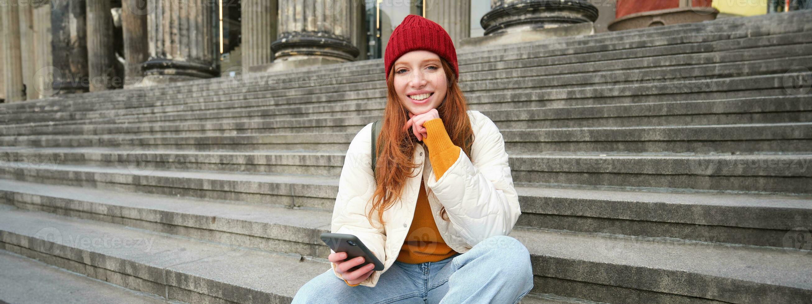 ritratto di giovane urbano ragazza nel rosso cappello, si siede su le scale vicino Museo, detiene mobile Telefono, si connette per pubblico Wi-Fi e surf rete, usi smartphone applicazioni foto