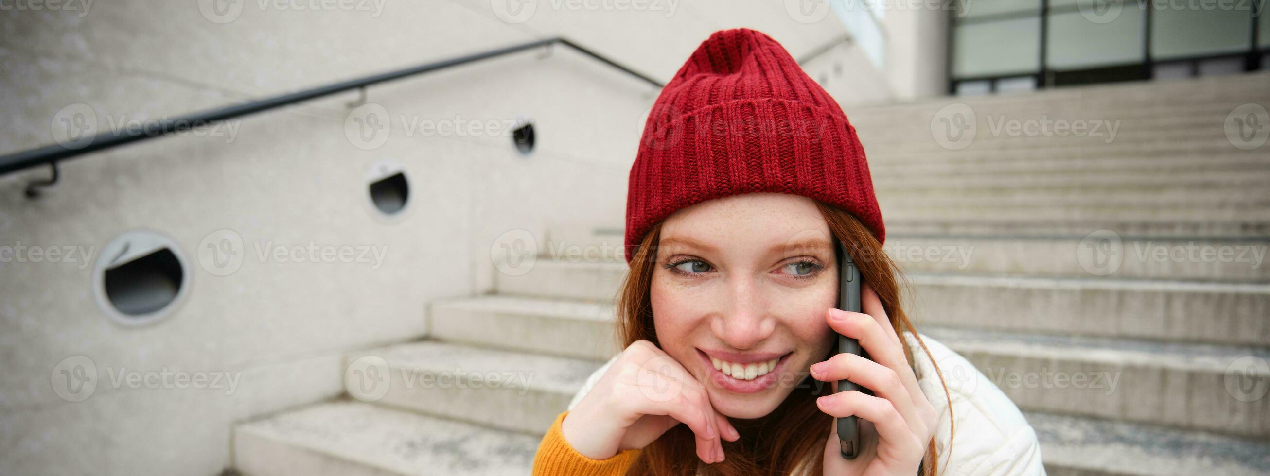 bellissimo sorridente testa Rossa femmina modello, si siede su strada e parla su mobile Telefono, usi smartphone App per chiamata all'estero, ridendo durante telefono conversazione foto