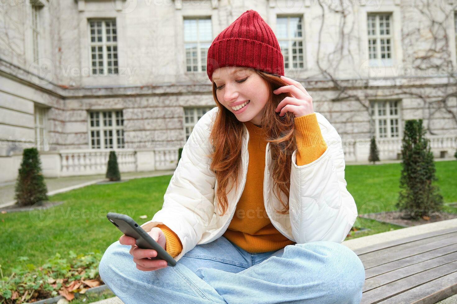giovane sorridente testa Rossa ragazza si siede su panchina e usi smartphone app, legge notizia in linea, orologi video su mobile Telefono mentre rilassato nel parco foto