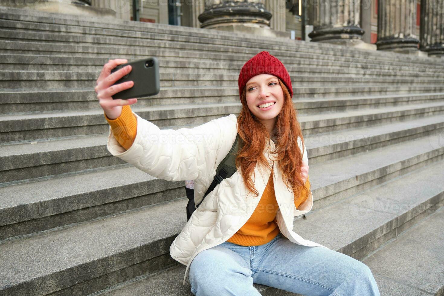 elegante giovane ragazza nel rosso cappello, prende fotografie su smartphone telecamera, fa autoscatto come lei si siede su le scale vicino Museo, in posa per foto con App filtro