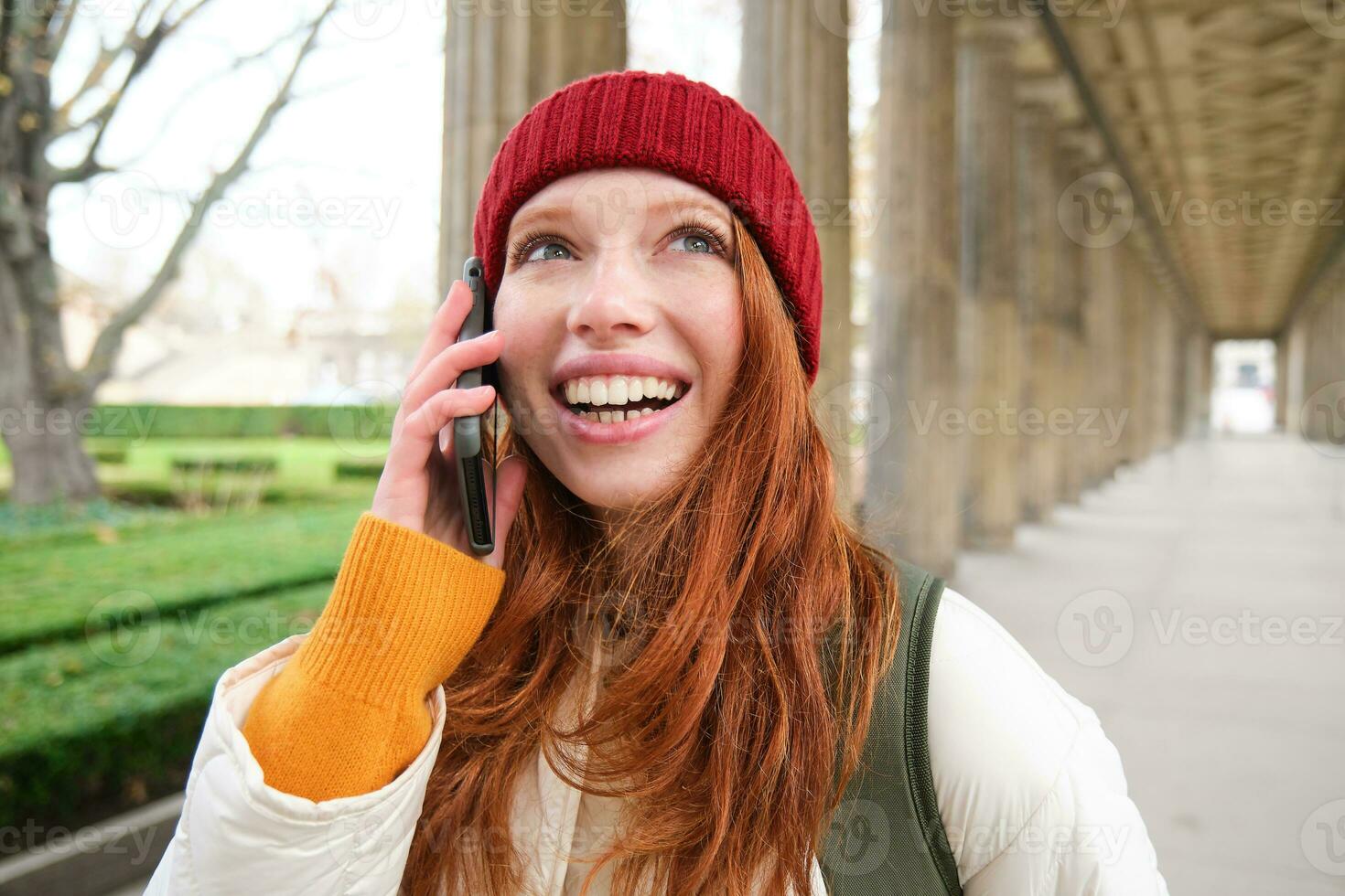 ritratto di testa Rossa europeo ragazza nel rosso cappello, fa un' Telefono chiamata, passeggiate nel città e parla per amico su smartphone foto