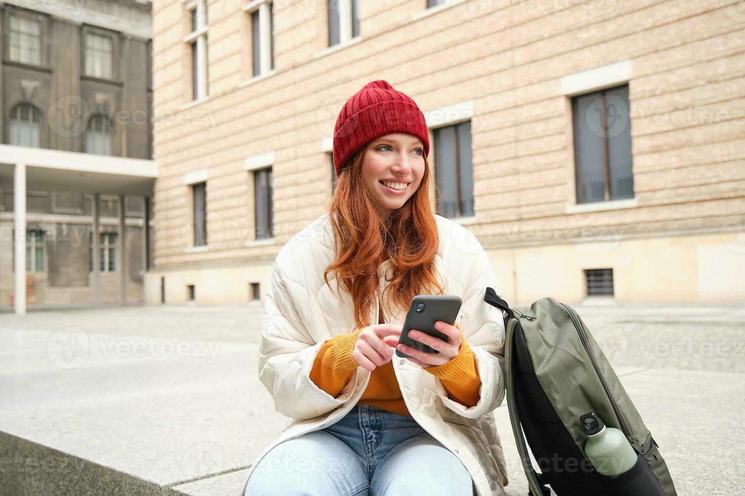 sociale media e le persone. giovane testa Rossa ragazza si siede su strada, usi mobile Telefono app, sembra su informazione nel Internet, detiene smartphone foto