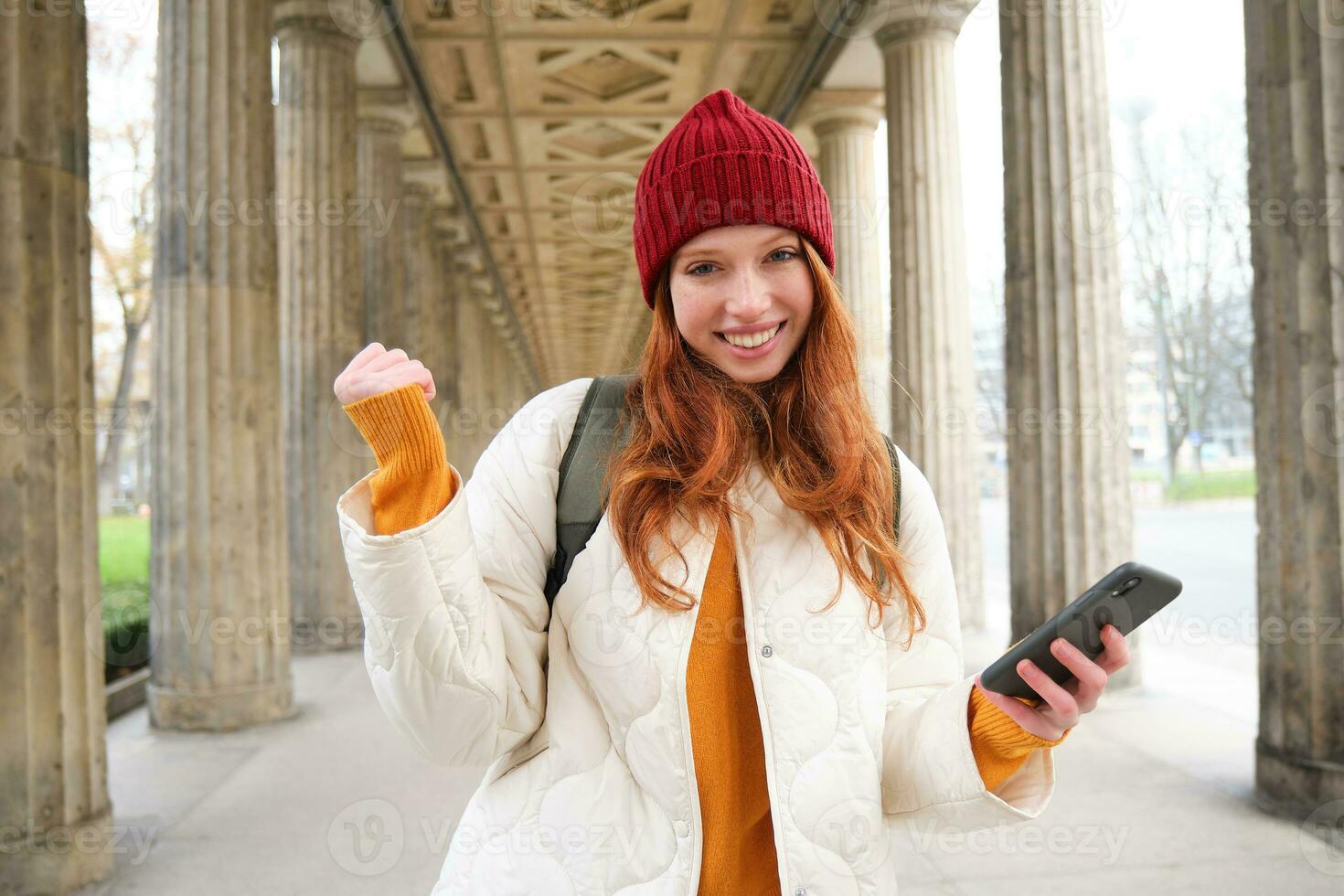 sorridente testa Rossa ragazza sembra emozionato, turista Tenere mobile Telefono e fa cazzotto pompa, entusiasta di sua viaggio, utilizzando smartphone su strada foto