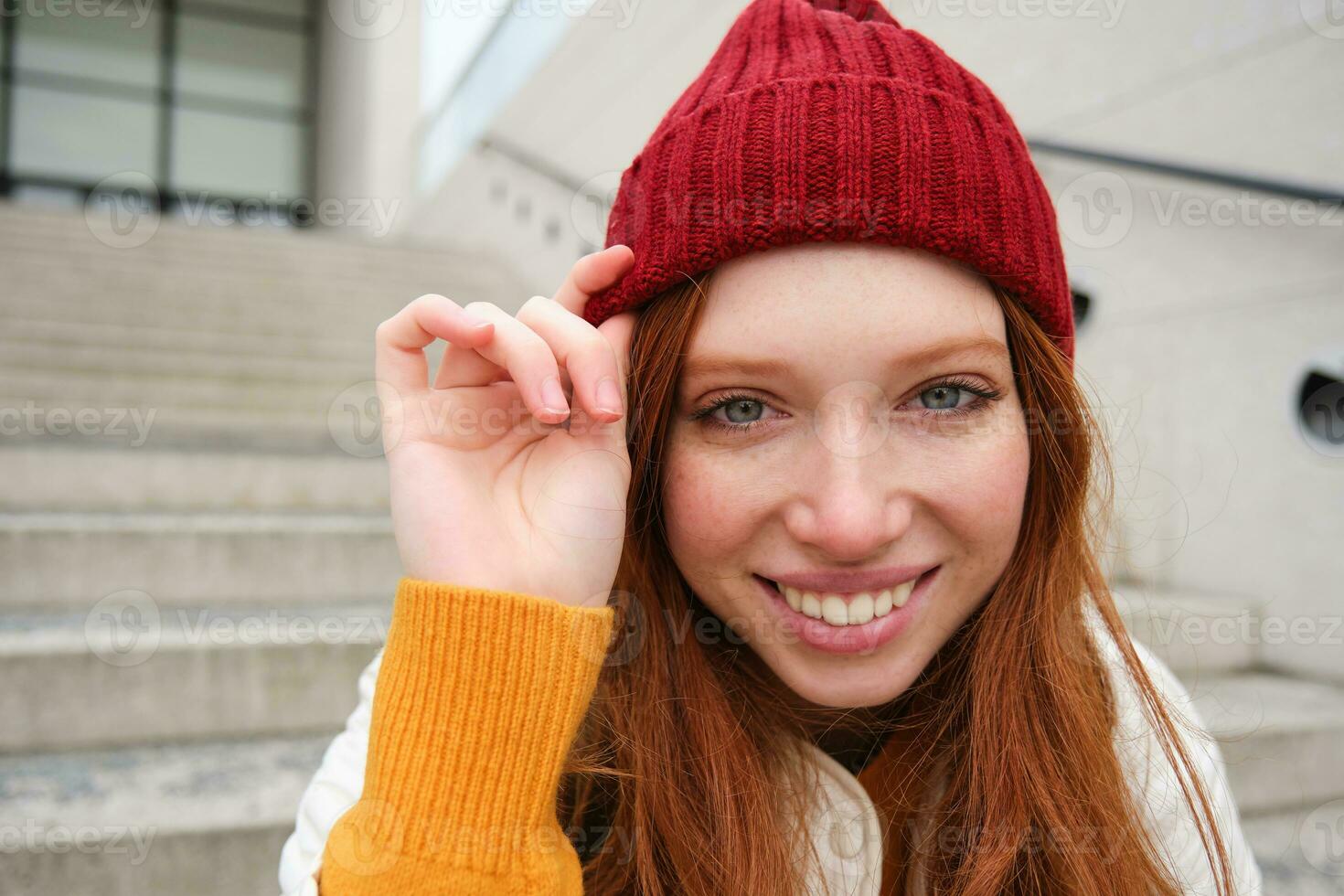 elegante testa Rossa ragazza nel caldo rosso cappello, sorridente rilassato, seduta con zaino su le scale vicino costruzione, aspetta per qualcuno all'aperto foto