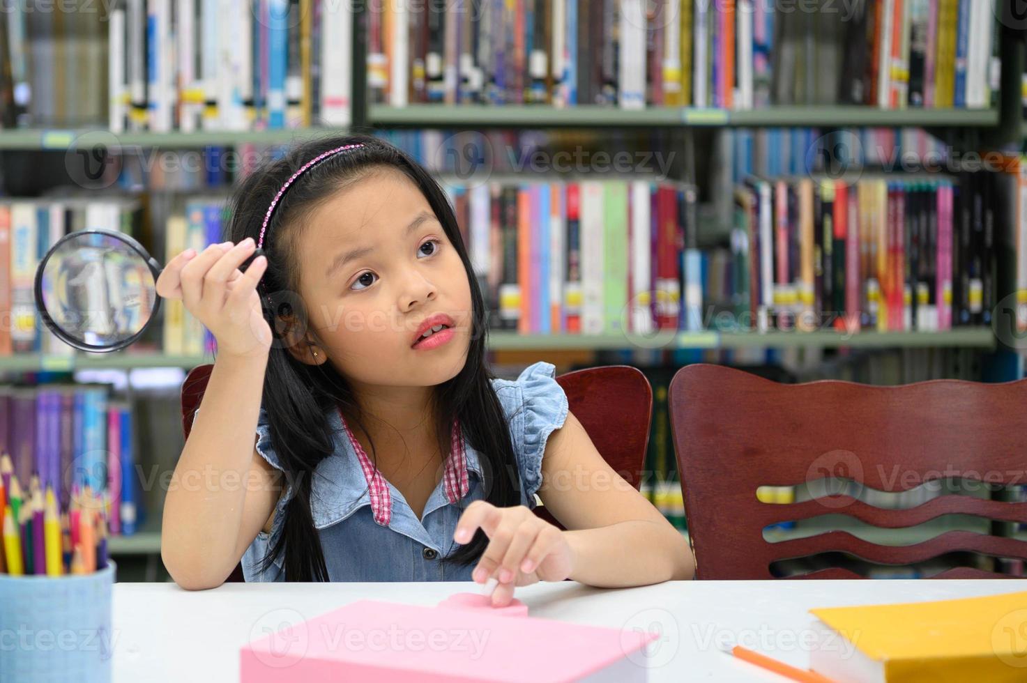 ragazza asiatica che tiene la lente d'ingrandimento e pensa in biblioteca a scuola. concetto di educazione e apprendimento foto