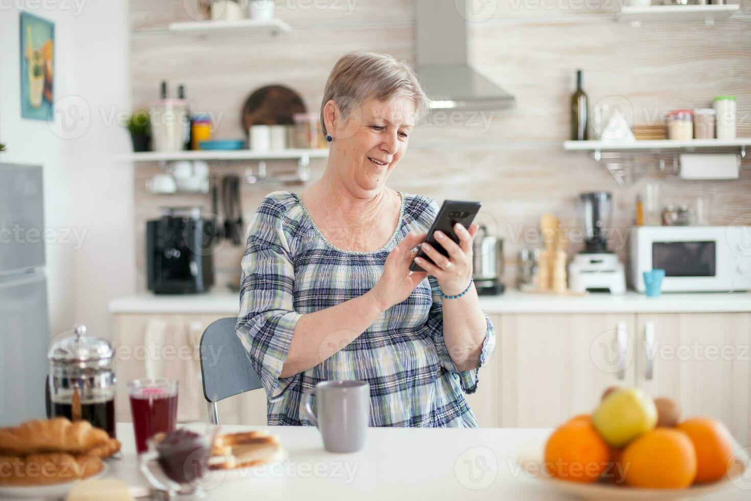 anziano donna utilizzando mobile aggeggio nel il cucina. autentico anziano persona utilizzando moderno smartphone Internet tecnologia. in linea comunicazione collegato per il mondo, anziano tempo libero tempo con aggeggio foto