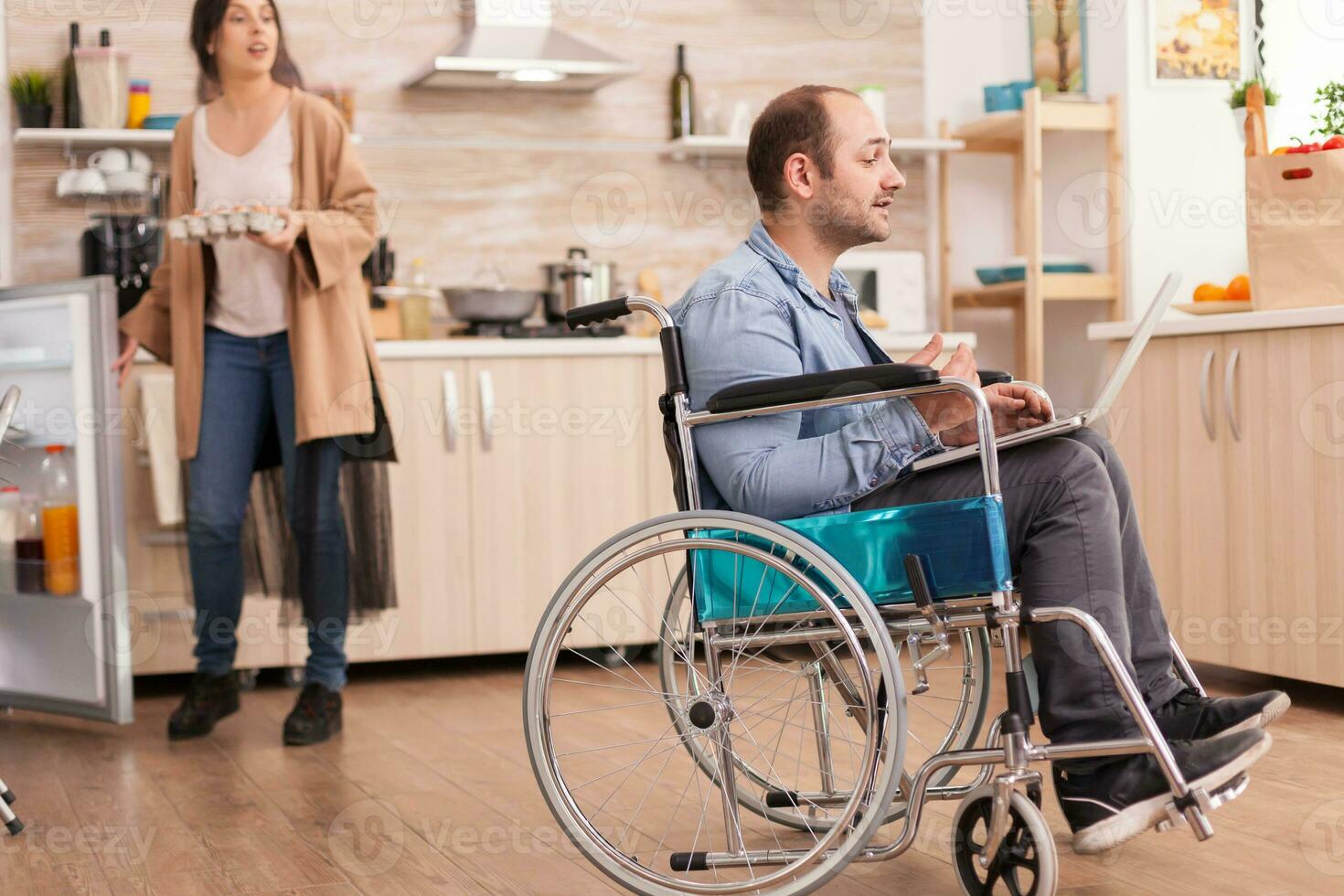 Disabilitato uomo nel sedia a rotelle durante video chiamata su il computer portatile nel cucina mentre è Tenere scatola di uova e Tenere frigorifero porta aprire. Disabilitato paralizzato portatori di handicap uomo con a piedi invalidità integrando dopo un incidente. foto