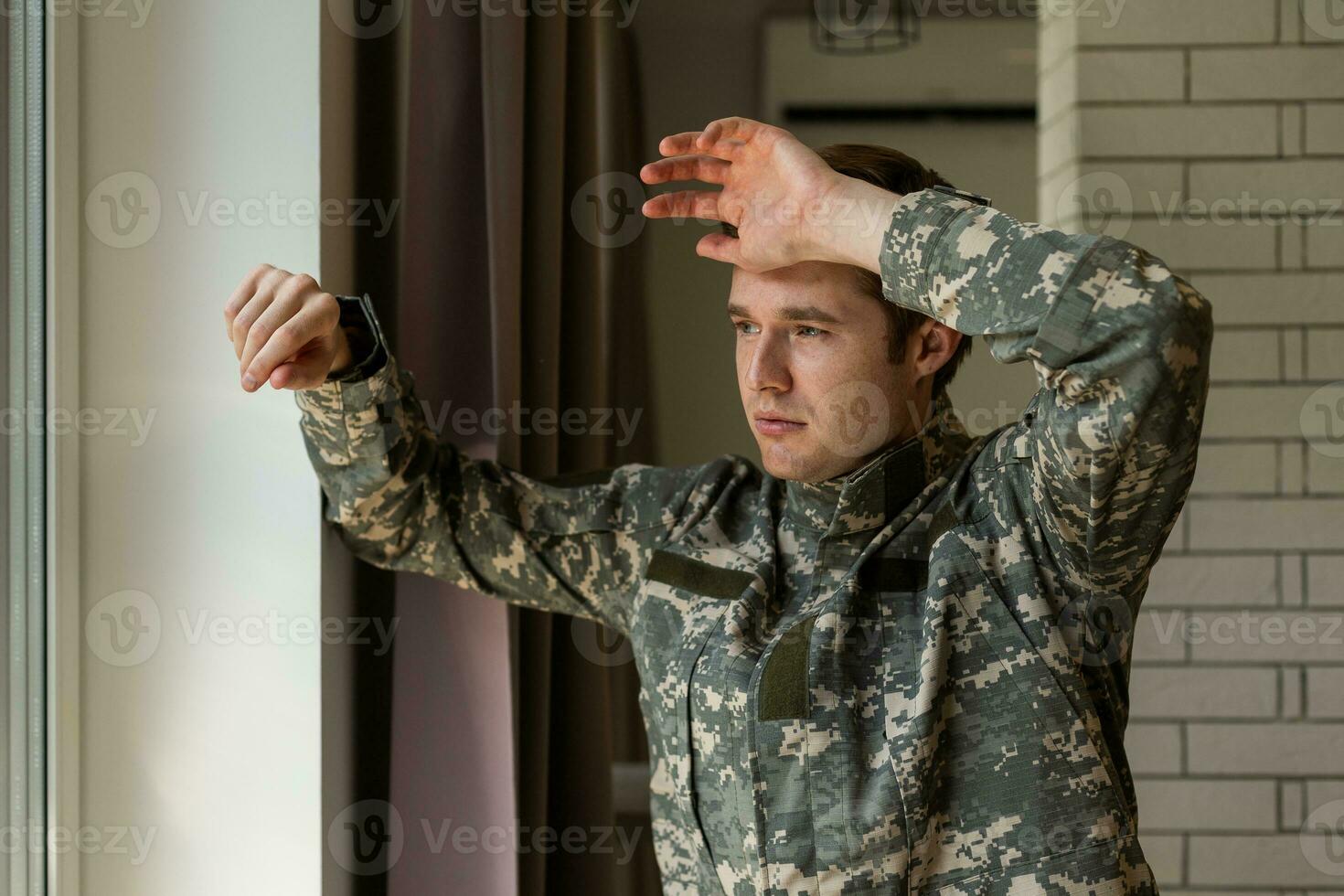 grave capelli corti giovane uomo nel militare uniforme soldato guardare attraverso il finestra, irritato soldato sofferenza a partire dal post traumatico sindrome dopo ritorno casa, copia spazio, avvicinamento. foto