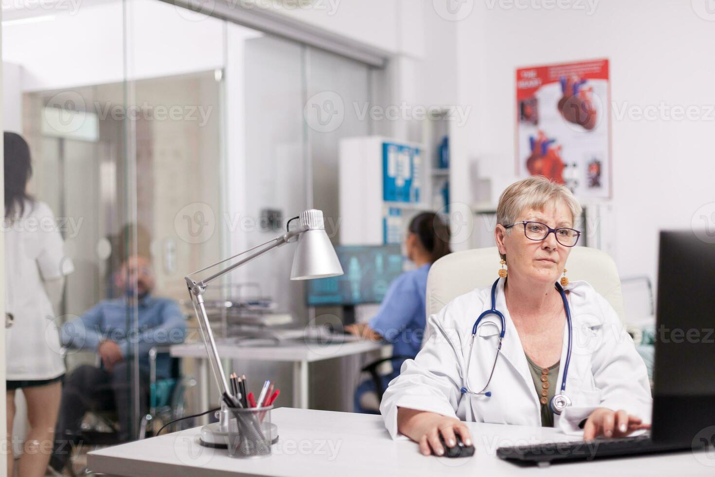 anziano donna medico Lavorando su computer nel ospedale ufficio mentre giovane medico è discutere con Disabilitato paziente nel sedia a rotelle su clinica corridoio e infermiera nel blu uniforme. foto