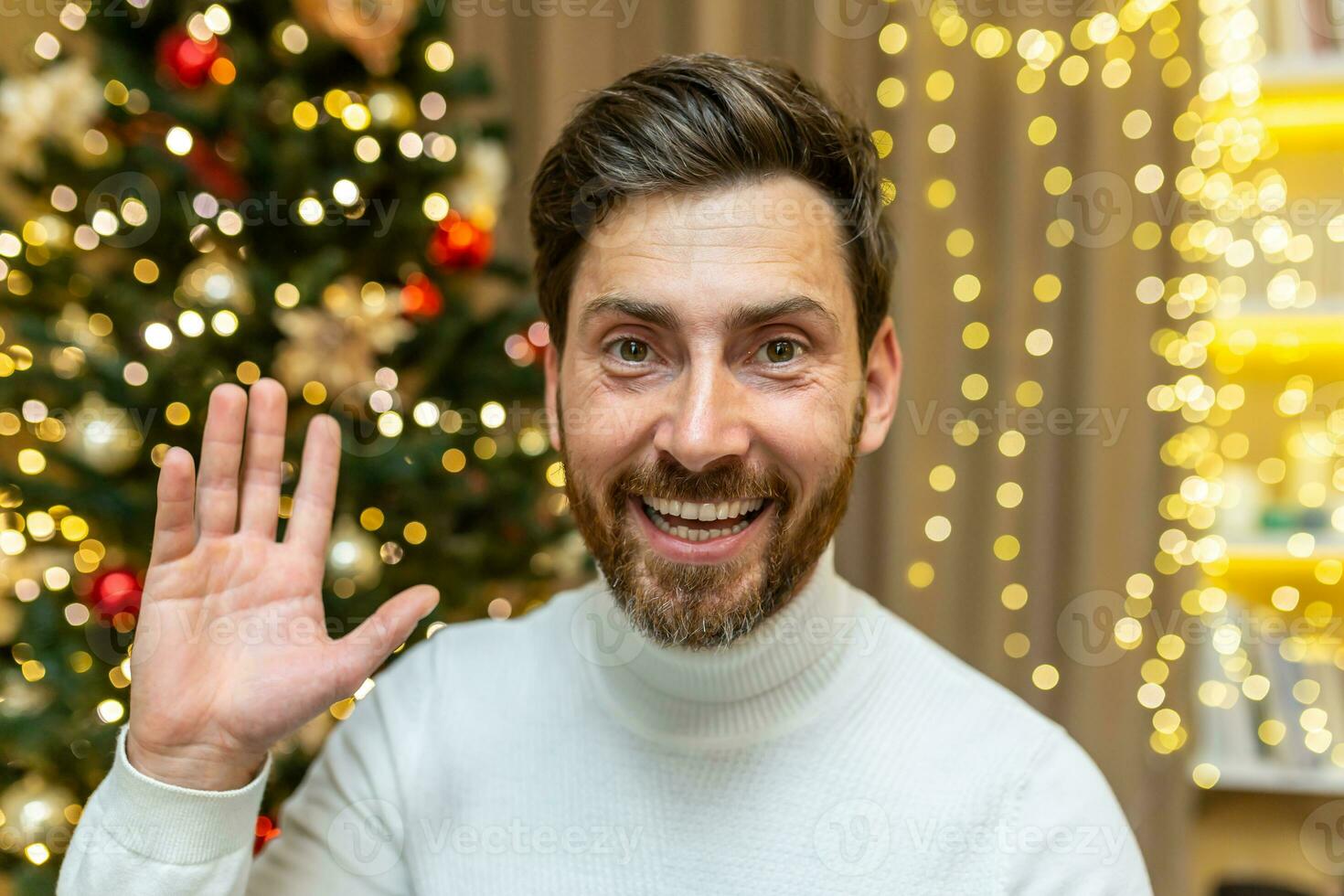 in linea saluti video chiamata, Natale uomo sembra in ragnatela telecamera e sorrisi e onde mentre seduta su divano a casa festeggiare nuovo anno vacanze. foto