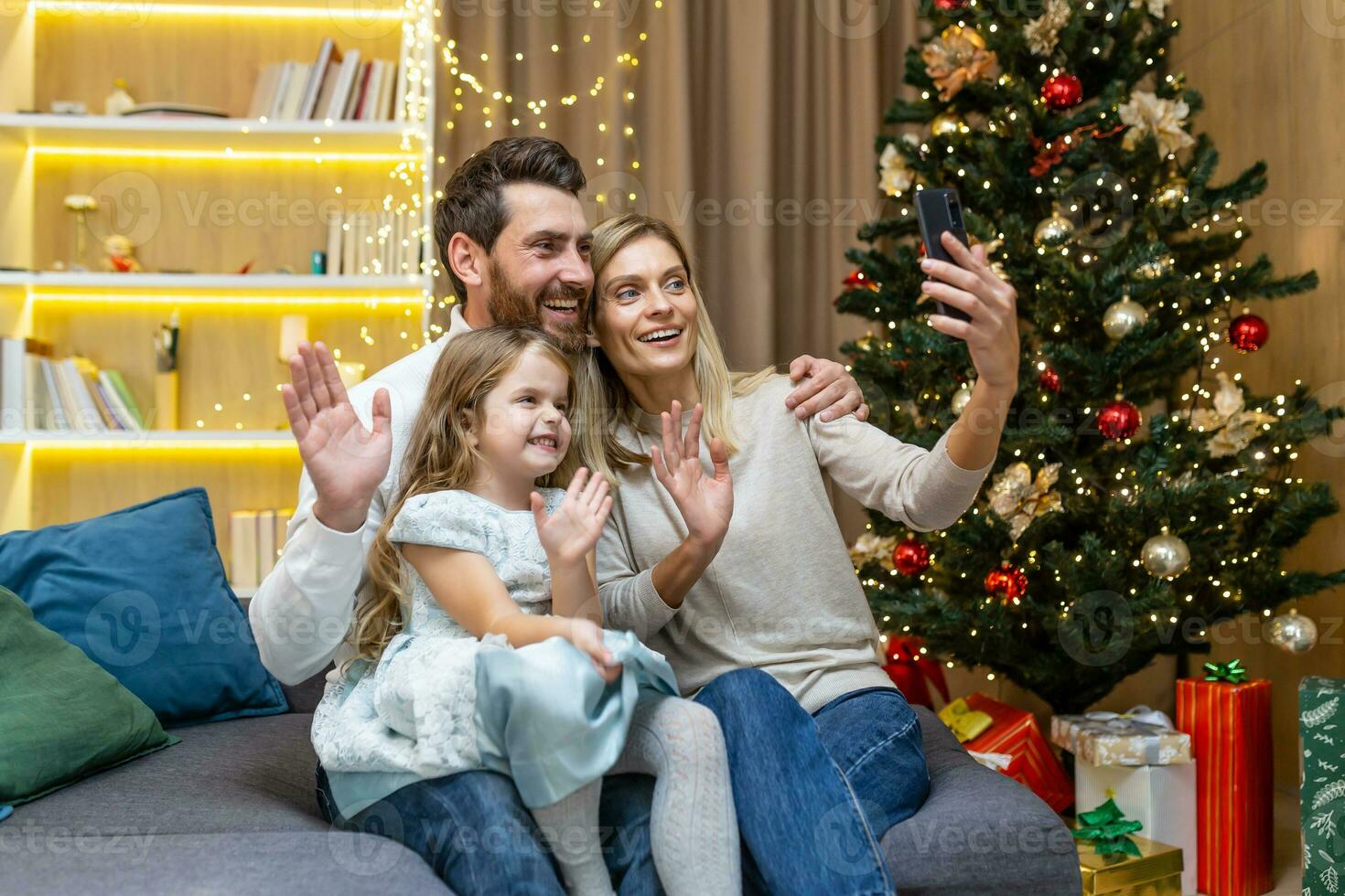 contento famiglia festeggiare nuovo anno e Natale, marito moglie e poco figlia seduta su divano nel vivente camera e parlando con parenti su video chiamata, utilizzando Telefono per a distanza comunicazione. foto