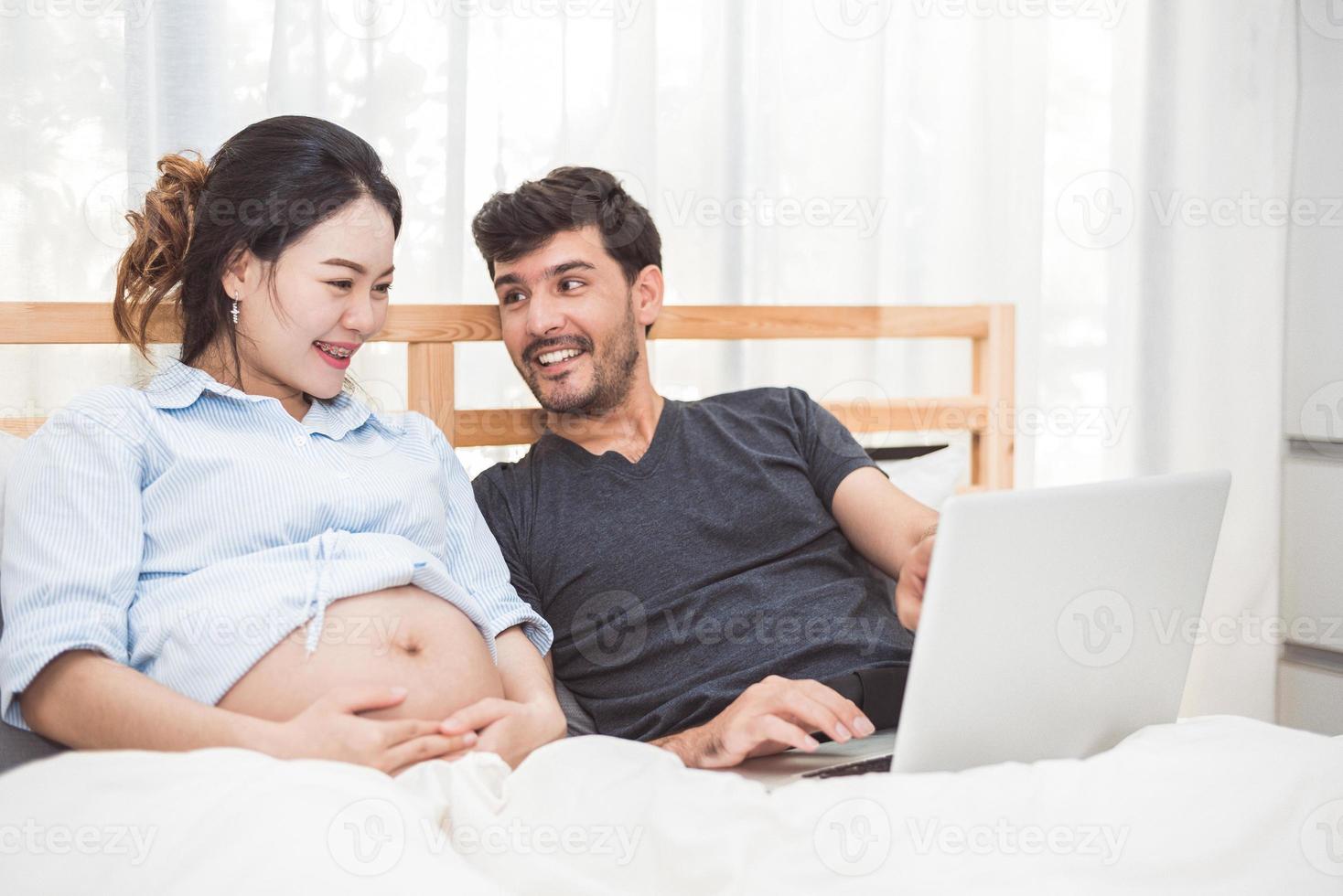 uomo felice e sua moglie incinta che usano il laptop per cercare articoli per neonati per preparare la genitorialità. coppia lifestyle famiglia e concetto di tecnologia. salute delle donne e tema medico foto