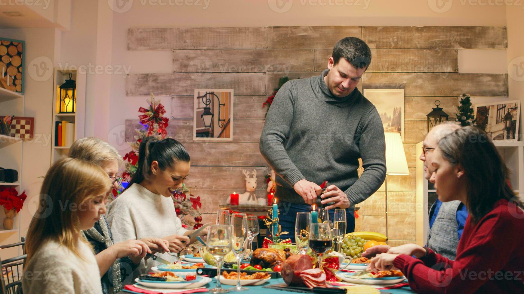 eccitato membri di il famiglia mentre il padre si apre un' bottiglia di vino a Natale riunione. tradizionale festivo Natale cena nel multigenerazionale famiglia. godendo natale pasto festa nel decorato camera. grande famiglia riunione foto