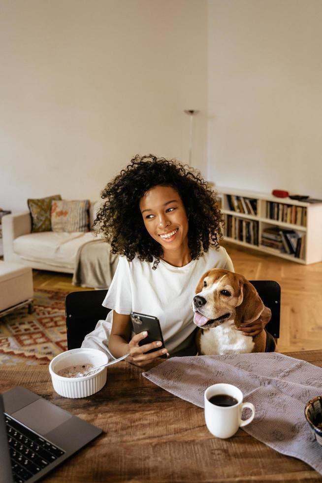 donna di colore che usa il cellulare e abbraccia il suo cane mentre fa colazione foto