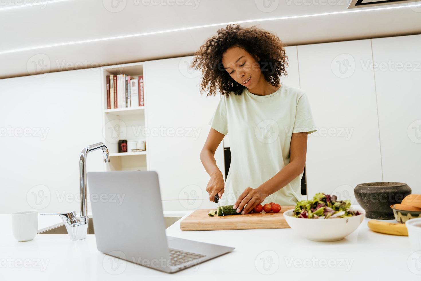 giovane donna nera che prepara insalata mentre usa il computer portatile in cucina foto