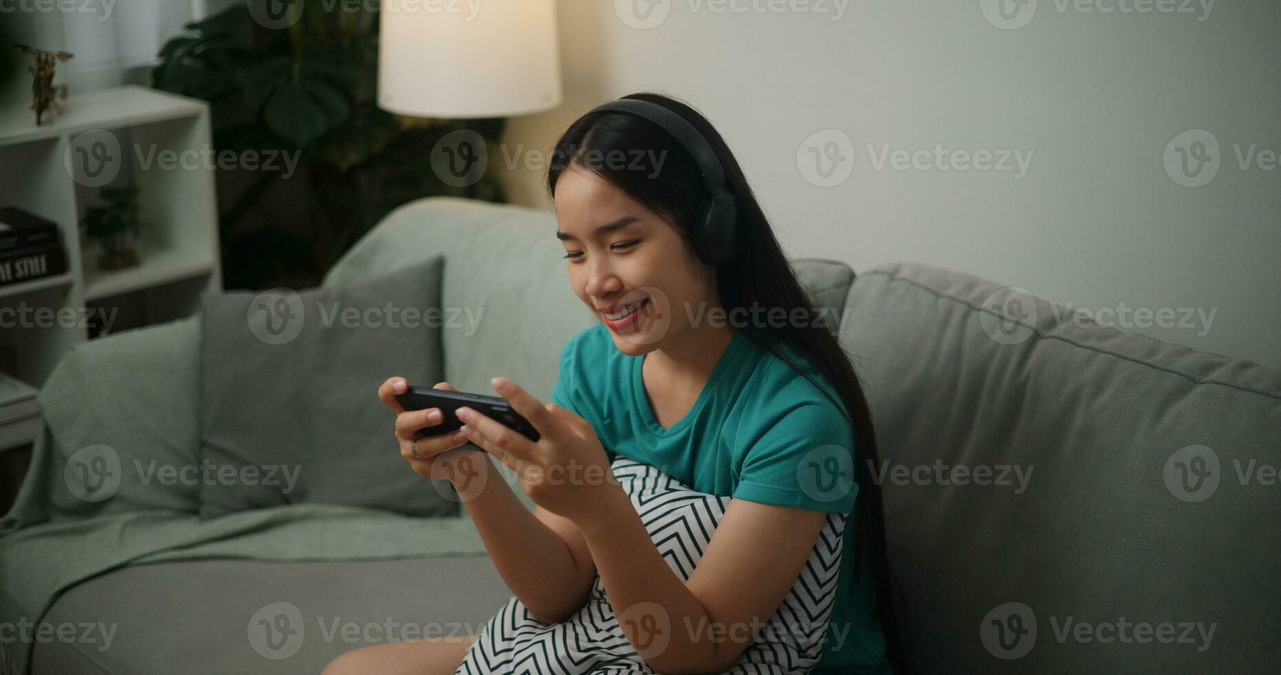 ritratto di giovane asiatico donna indossare bicchieri e cuffie gode giocando in linea esport Giochi su smartphone seduta su divano nel il vivente camera a casa, giocatore stile di vita concetto. foto