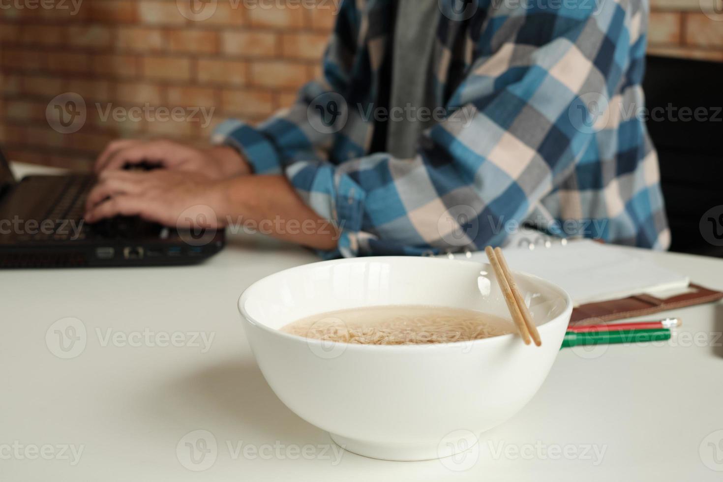 lavoratore di sesso maschile tailandese impegnato a lavorare con il laptop, usa le bacchette per mangiare frettolosamente noodles istantanei durante la pausa pranzo in ufficio, perché veloce, gustoso ed economico. nel tempo fast food asiatico, stile di vita malsano. foto