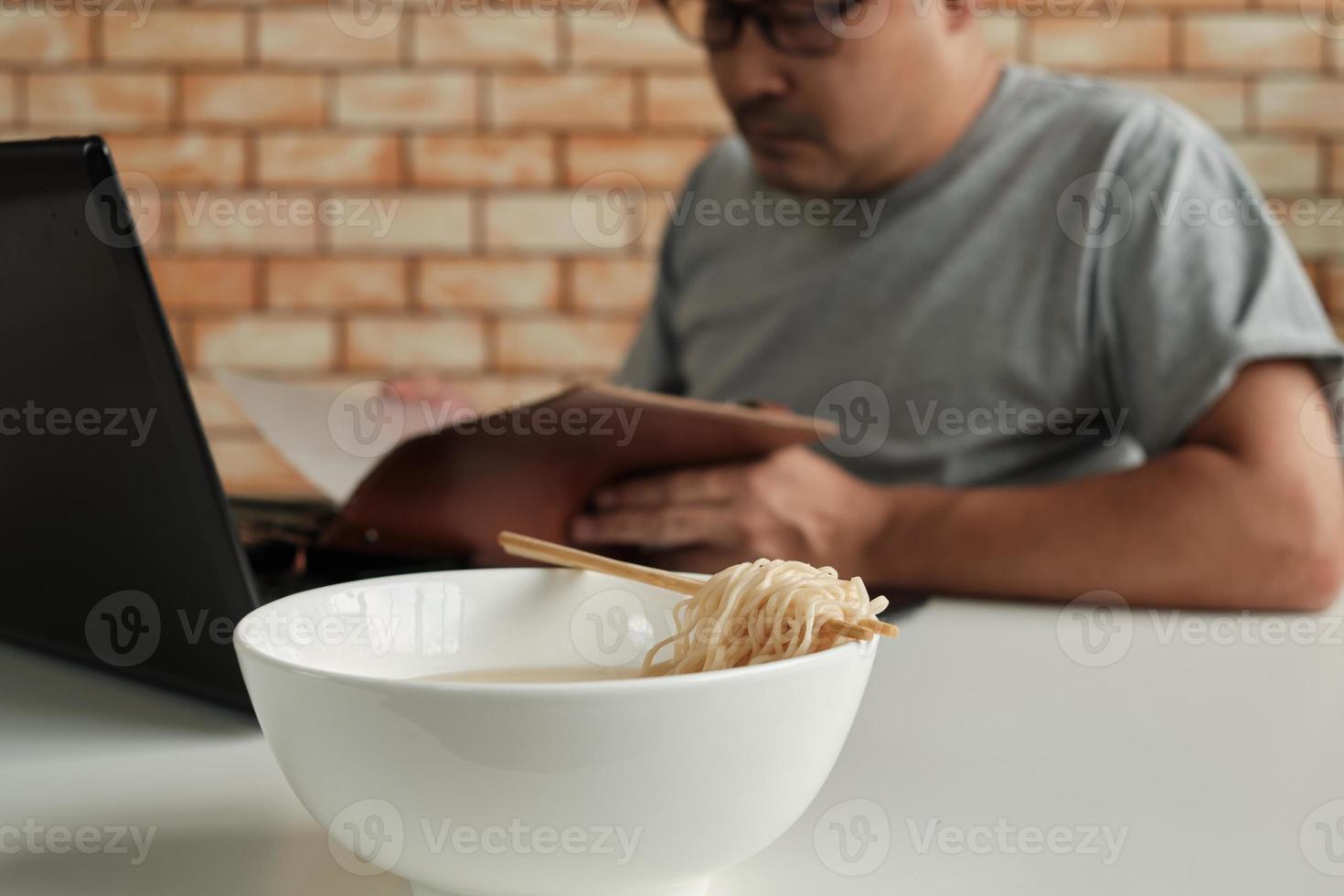 lavoratore di sesso maschile tailandese impegnato a lavorare con il laptop, usa le bacchette per mangiare frettolosamente noodles istantanei durante la pausa pranzo in ufficio, perché veloce, gustoso ed economico. nel tempo fast food asiatico, stile di vita malsano. foto