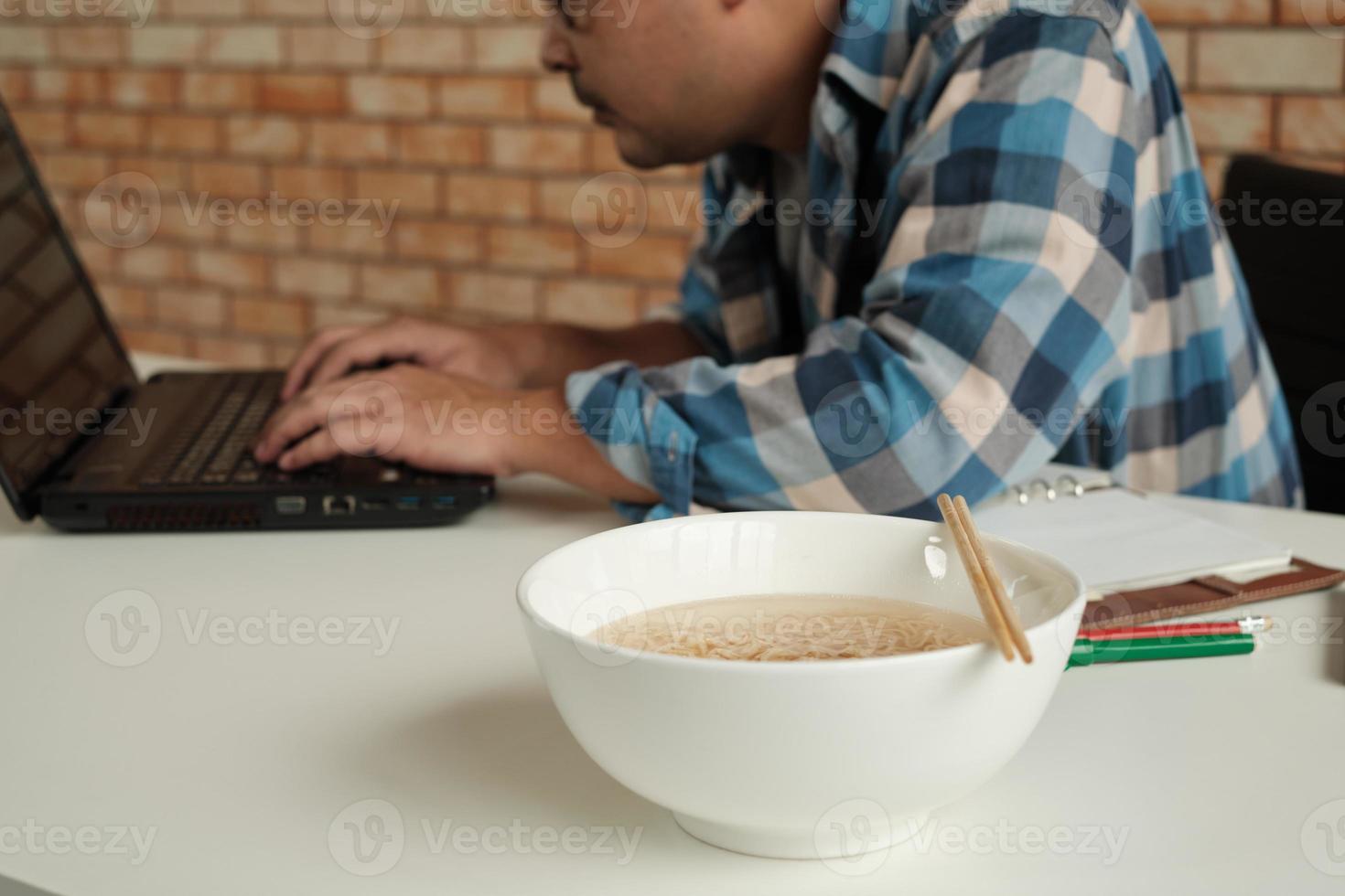 lavoratore di sesso maschile tailandese impegnato a lavorare con il laptop, usa le bacchette per mangiare frettolosamente noodles istantanei durante la pausa pranzo in ufficio, perché veloce, gustoso ed economico. nel tempo fast food asiatico, stile di vita malsano. foto