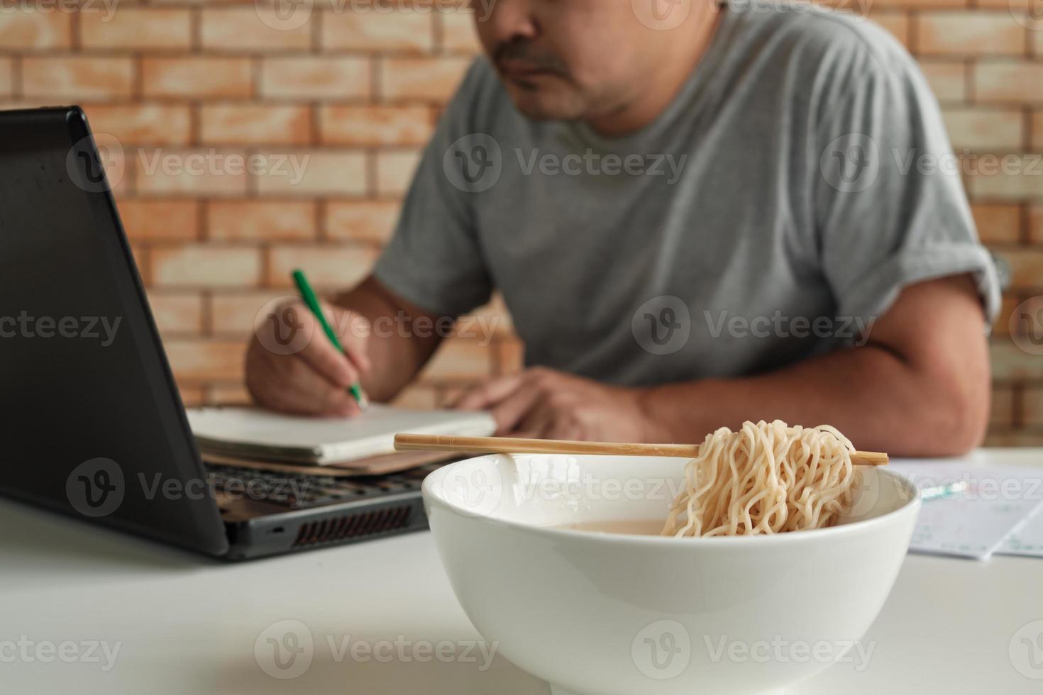 lavoratore di sesso maschile tailandese impegnato a lavorare con il laptop, usa le bacchette per mangiare frettolosamente noodles istantanei durante la pausa pranzo in ufficio, perché veloce, gustoso ed economico. nel tempo fast food asiatico, stile di vita malsano. foto