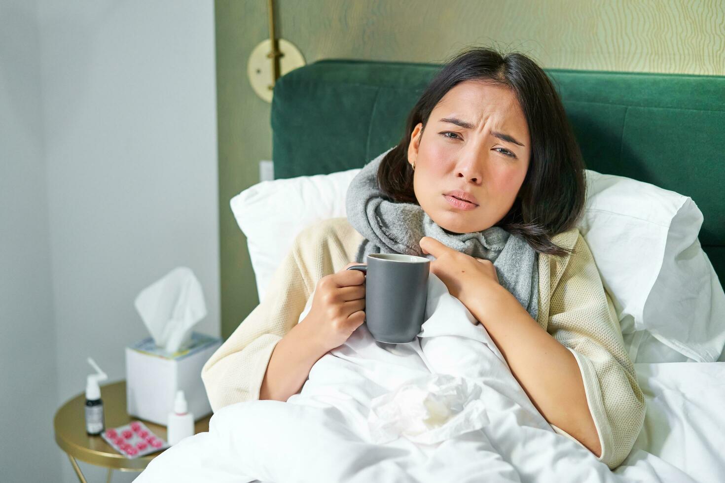 ritratto di adagiarsi asiatico ragazza nel letto, sensazione malato, attraente freddo e soggiorno a casa, guardare malsano, potabile caldo tè foto