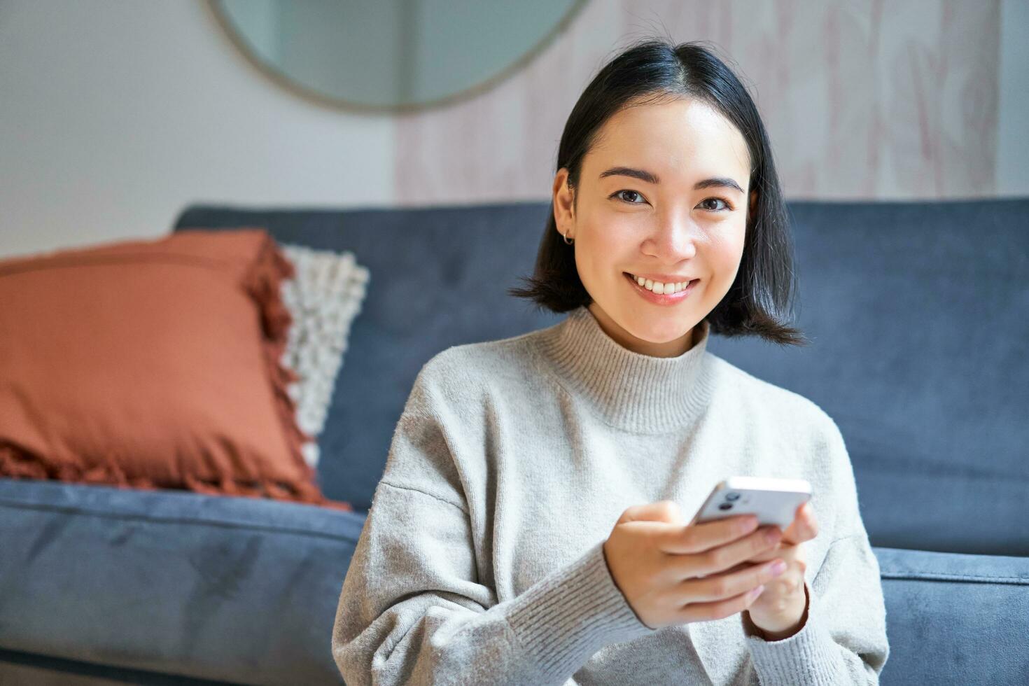 persone e tecnologia. sorridente asiatico donna seduta a casa, utilizzando sua mobile Telefono, digitando Messaggio, sbirciando Internet o shopping in linea a partire dal smartphone App foto