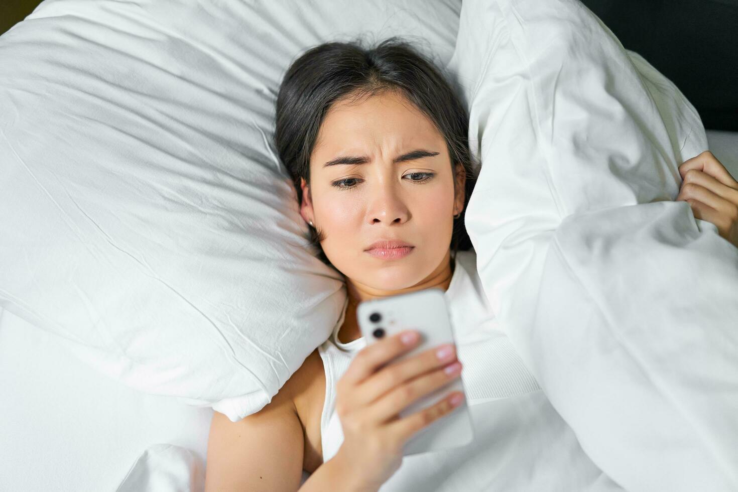 vicino su ritratto di asiatico ragazza dire bugie nel letto, guardare a smartphone ha riguardato, veglia su in ritardo e fissando a sua allarme orologio su mobile Telefono foto