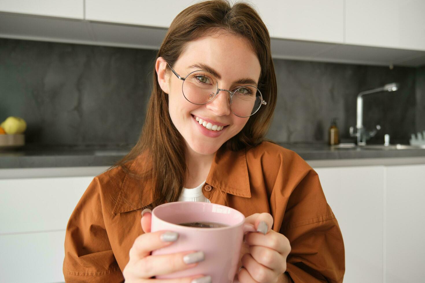 vicino su ritratto di bellissimo donna nel occhiali, bevande Tè, riscalda sua mani con caldo tazza, sorridente e guardare contento a telecamera, seduta nel cucina foto