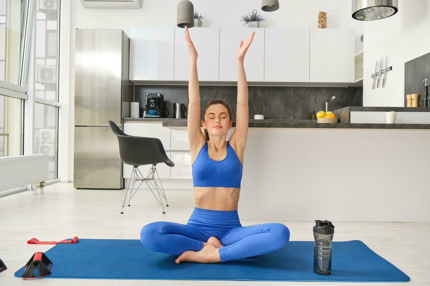 sorridente bellissimo ragazza fare yoga nel sua vivente camera, raccolta mani su e in profondità respirazione, pratica consapevolezza e rilassamento la pratica, seduta con gambe attraversato su gomma da cancellare stuoia foto