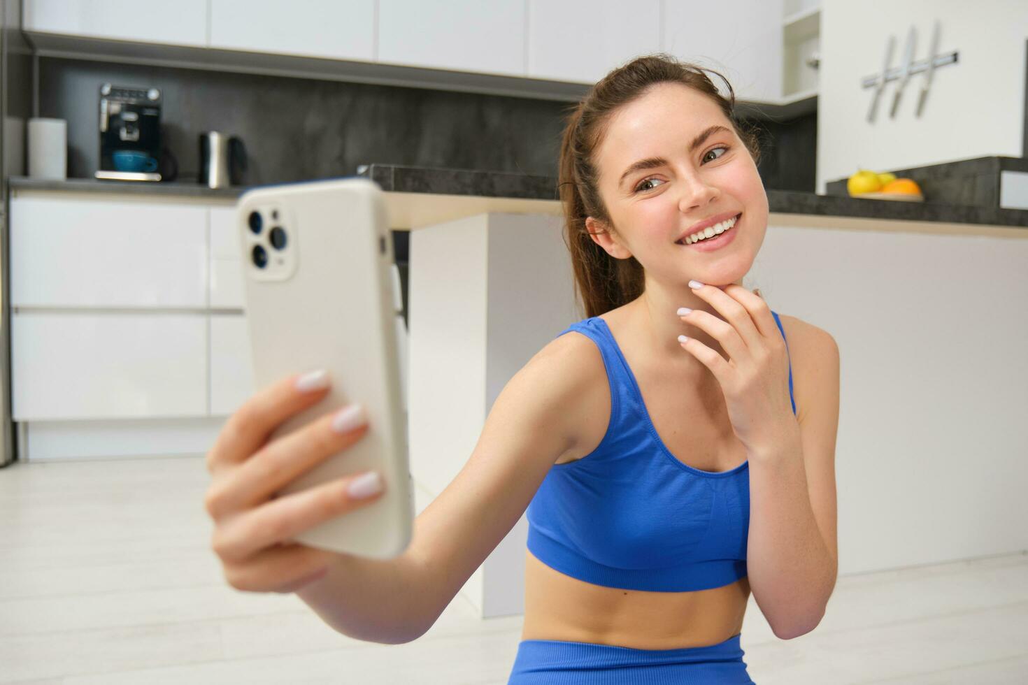 ritratto di giovane fitness ragazza, blogger record sua allenarsi a partire dal casa, pose per autoscatto su smartphone, fa yoga su telecamera, si siede nel blu reggiseno sportivo e ghette foto