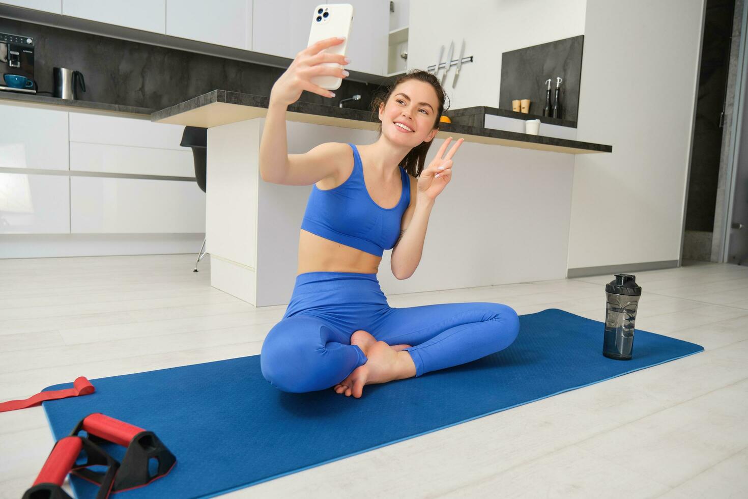 giovane sorridente donna, fittezza ragazza fare allenarsi a partire dal casa, si siede su yoga stuoia con smartphone, assunzione autoscatto, record sport video blog in casa foto
