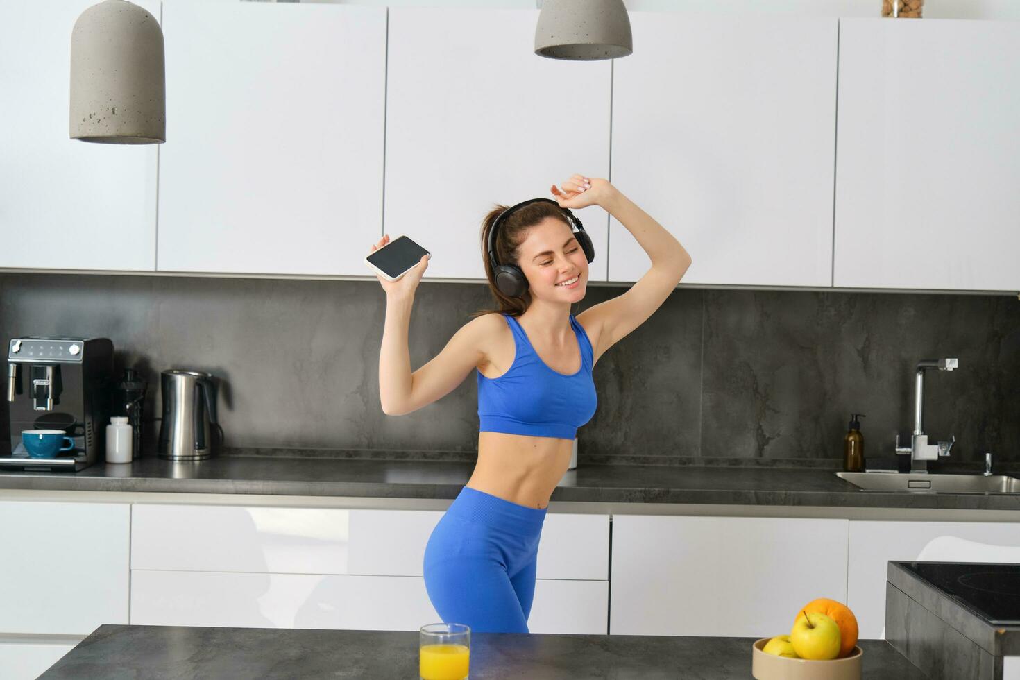 ritratto di bellissimo fitness blogger, donna nel cuffia, ascoltando musica e danza nel cucina, indossare blu ghette e reggiseno sportivo foto