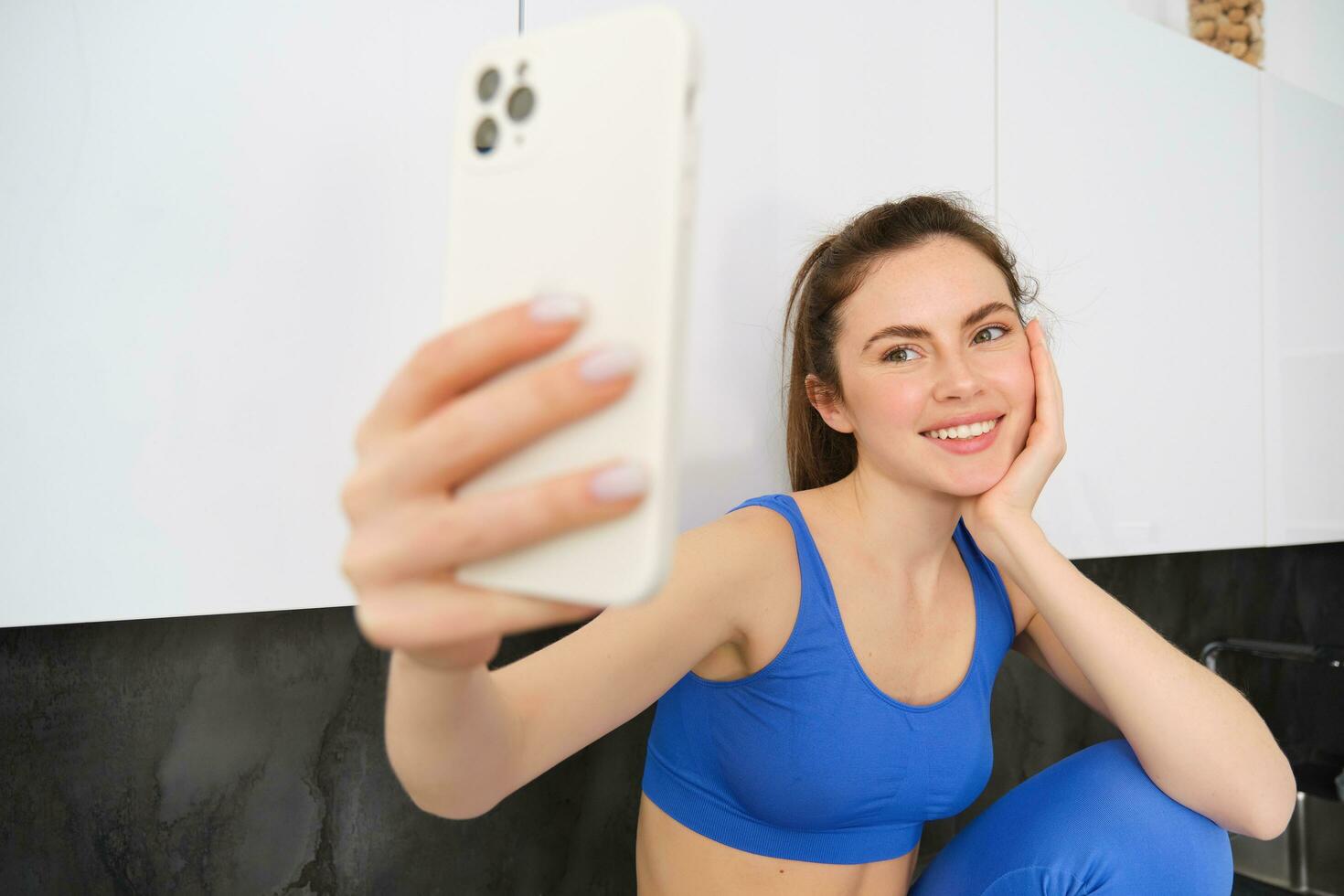 ritratto di fitness ragazza in posa per foto, assunzione autoscatto su smartphone app, seduta nel cucina, indossare abbigliamento sportivo foto