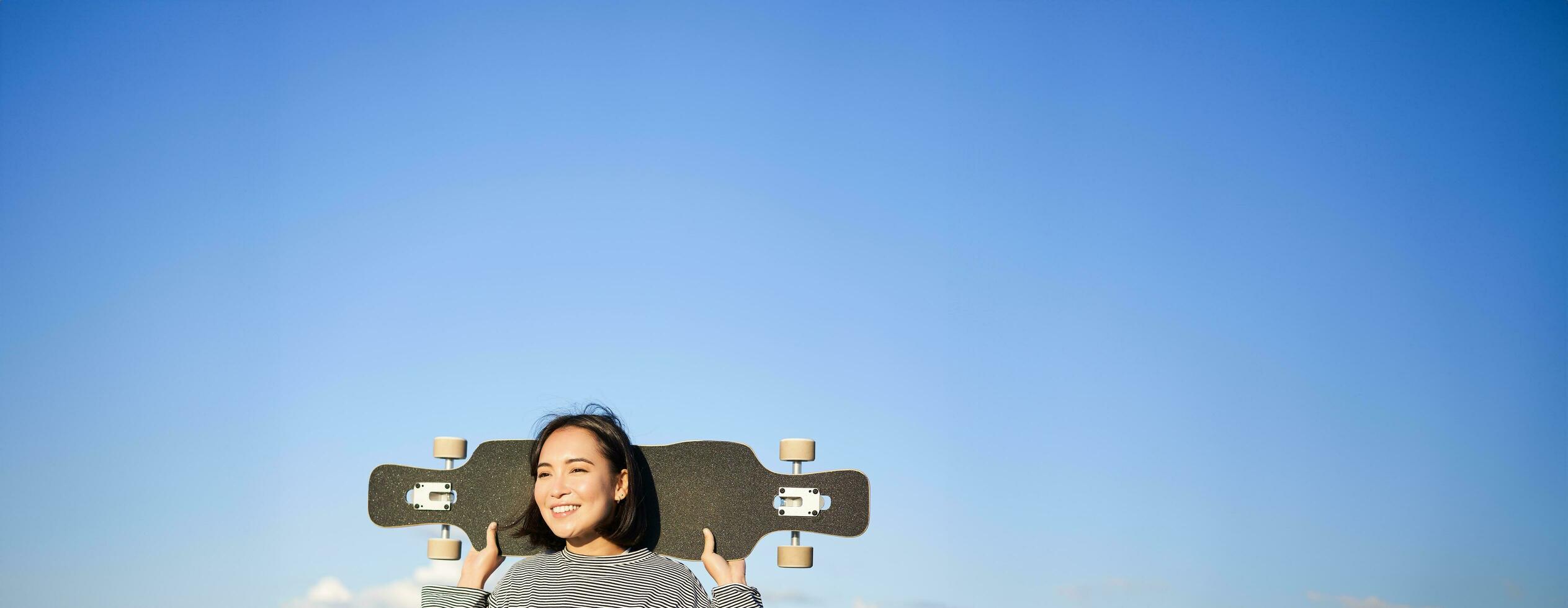 verticale tiro di spensierato asiatico ragazza con longboard. giovane donna pattinatore Tenere incrociatore su sua le spalle e a piedi su strada, andare con lo skateboard foto