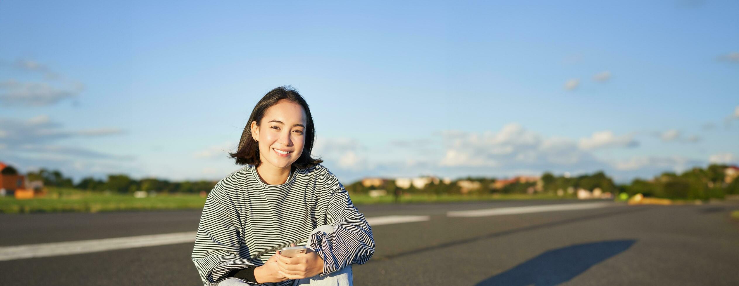 verticale tiro di asiatico donna seduta su skateboard su strada, Tenere smartphone app. pattinatore ragazza pattini su longboard, utilizzando mobile Telefono foto