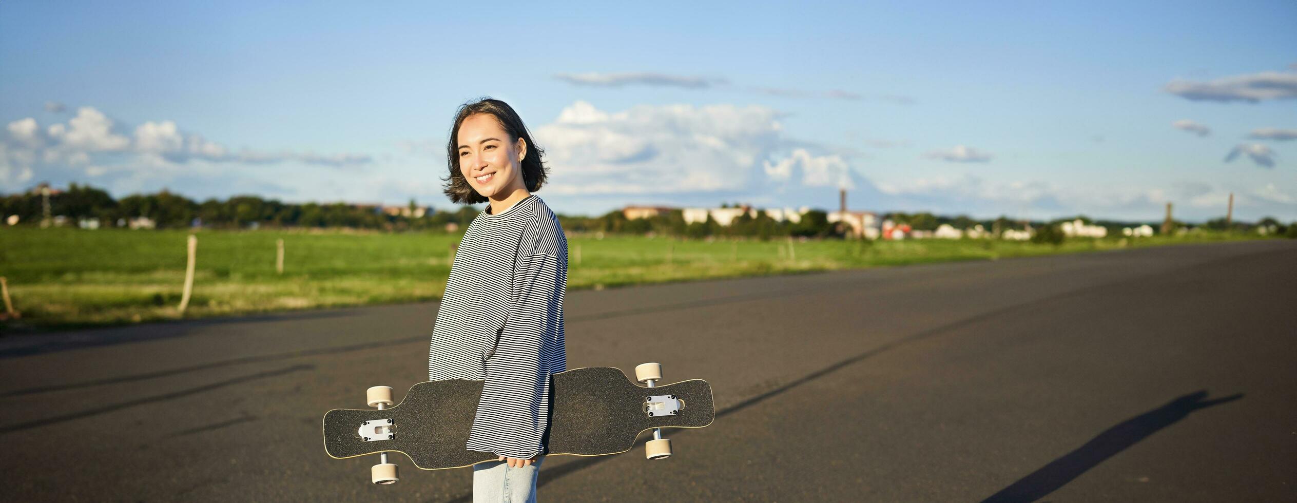 verticale tiro di spensierato asiatico ragazza con longboard. giovane donna pattinatore Tenere incrociatore su sua le spalle e a piedi su strada, andare con lo skateboard foto