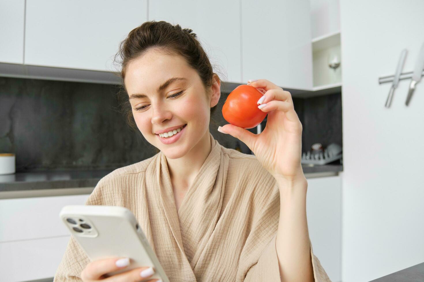 Immagine di sorridente bellissimo donna, si siede nel cucina, detiene smartphone e pomodoro, sembra felice, ordini drogheria consegna per sua casa, utilizzando mobile Telefono app, acquisto in linea verdure per sua ricetta foto