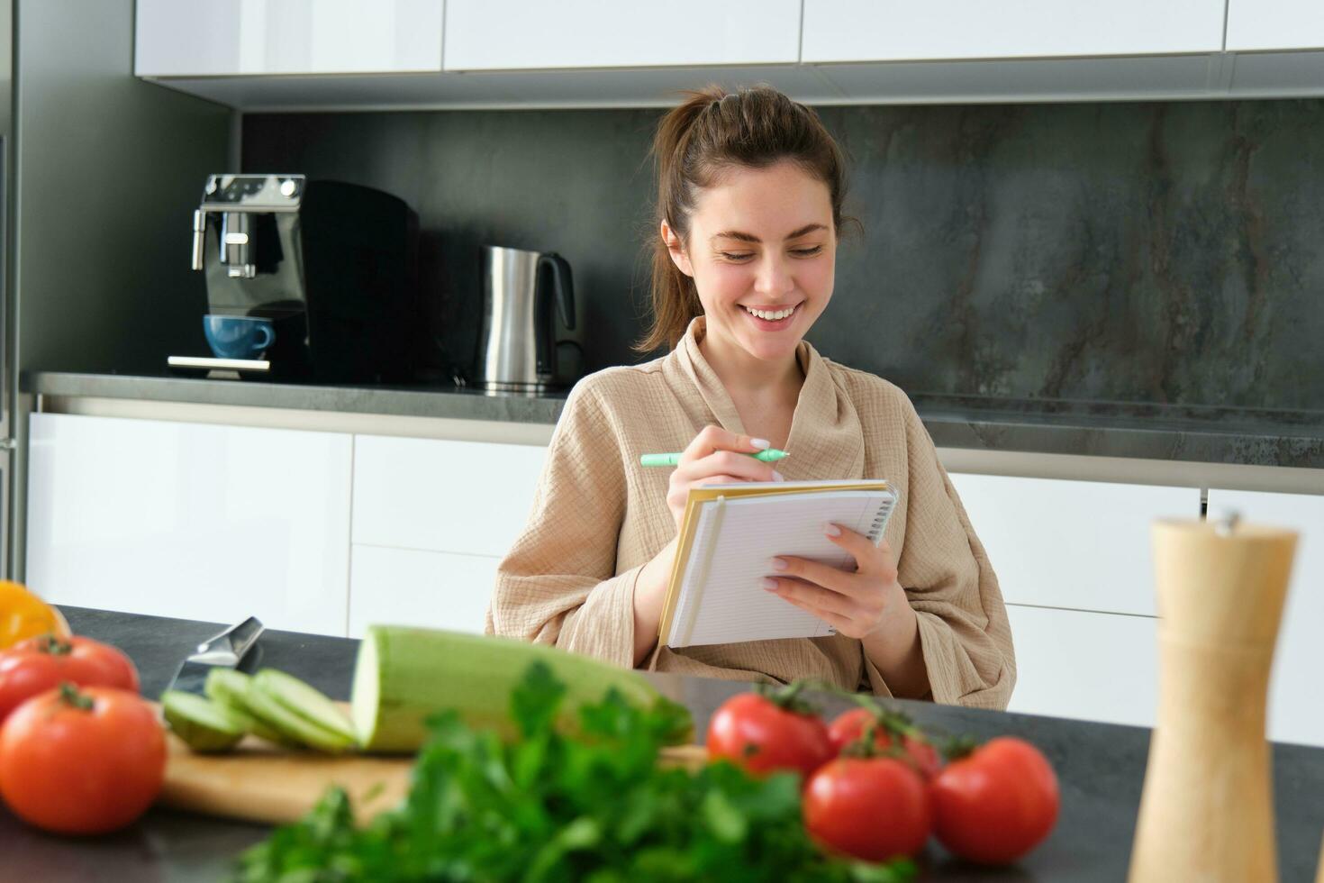 ritratto di Bellissima, sorridente giovane donna fabbricazione elenco di pasti, scrittura giù ricetta, seduta nel il cucina con la verdura, fare Casa commissioni foto