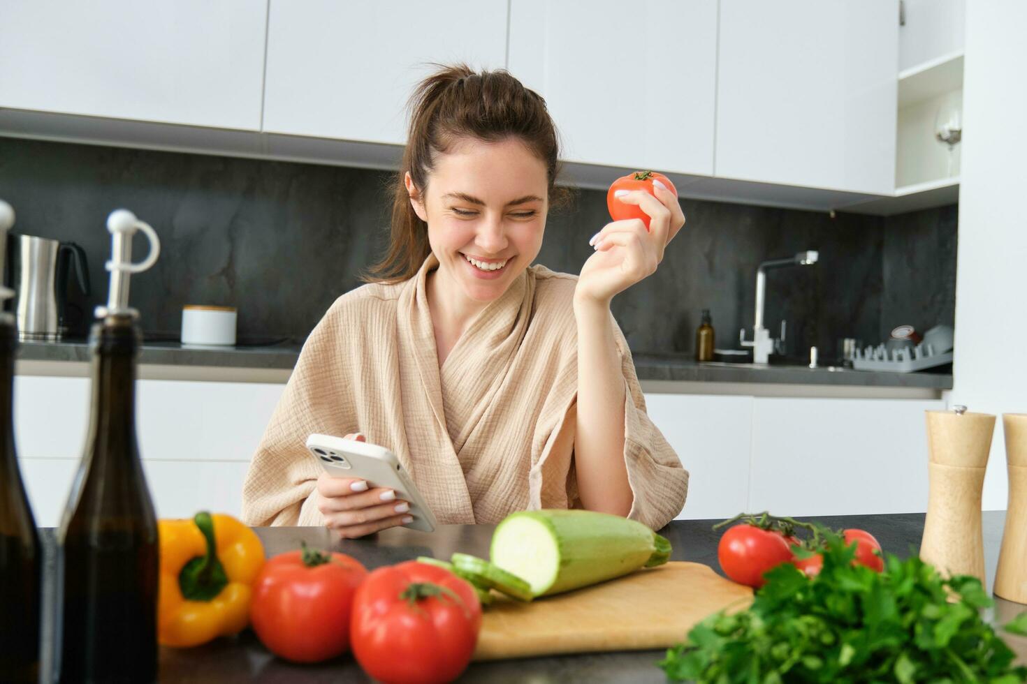 giovane donna ordini drogheria su mobile app. ragazza nel accappatoio si siede nel il cucina con la verdura, guardare per ricetta per cucinare cena, utilizzando smartphone applicazione foto