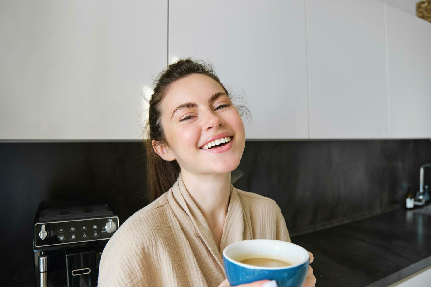 stile di vita concetto. ritratto di contento brunetta donna nel accappatoio, potabile caffè nel il cucina, avendo mattina cuppa e sorridente foto