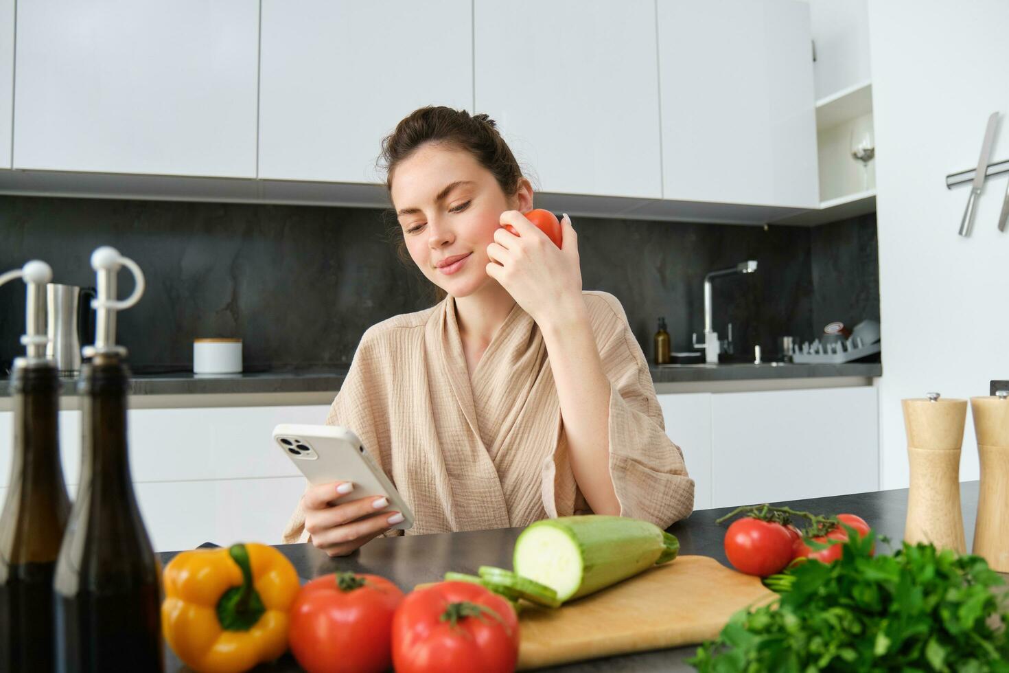 giovane donna ordini drogheria su mobile app. ragazza nel accappatoio si siede nel il cucina con la verdura, guardare per ricetta per cucinare cena, utilizzando smartphone applicazione foto