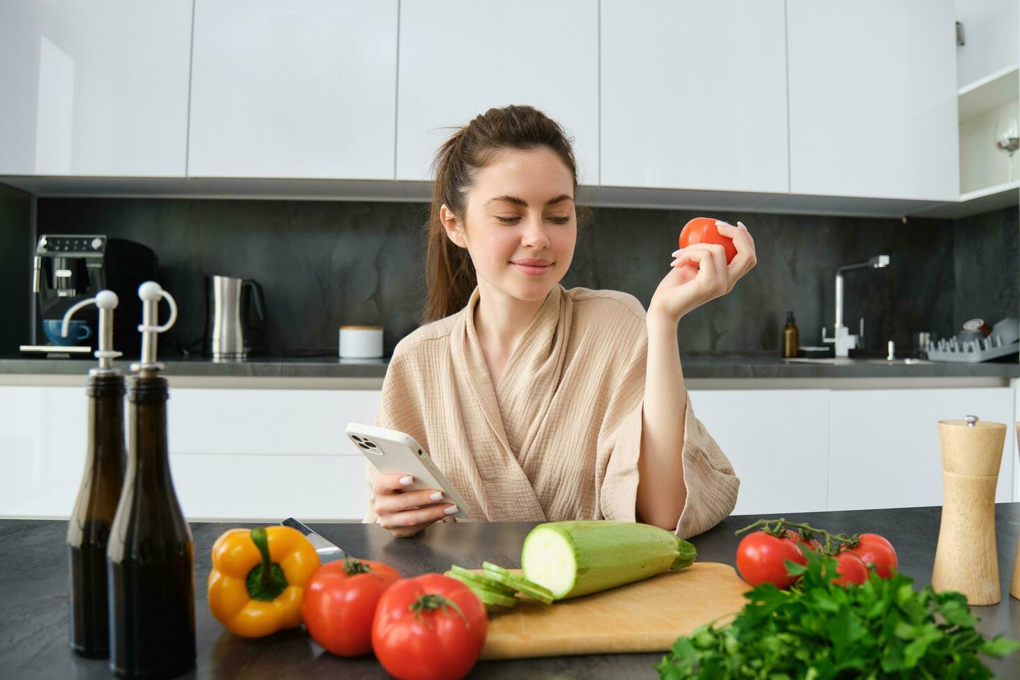 giovane donna ordini drogheria su mobile app. ragazza nel accappatoio si siede nel il cucina con la verdura, guardare per ricetta per cucinare cena, utilizzando smartphone applicazione foto