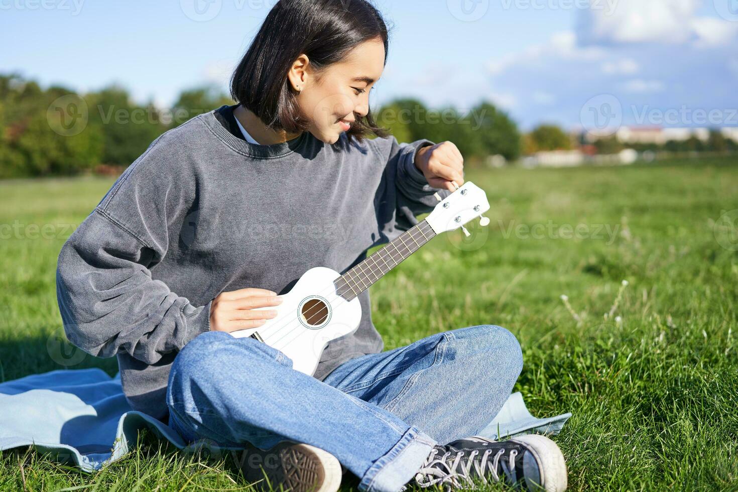 cantando asiatico ragazza giocando ukulele su erba, seduta su coperta nel parco, rilassante all'aperto su soleggiato giorno foto