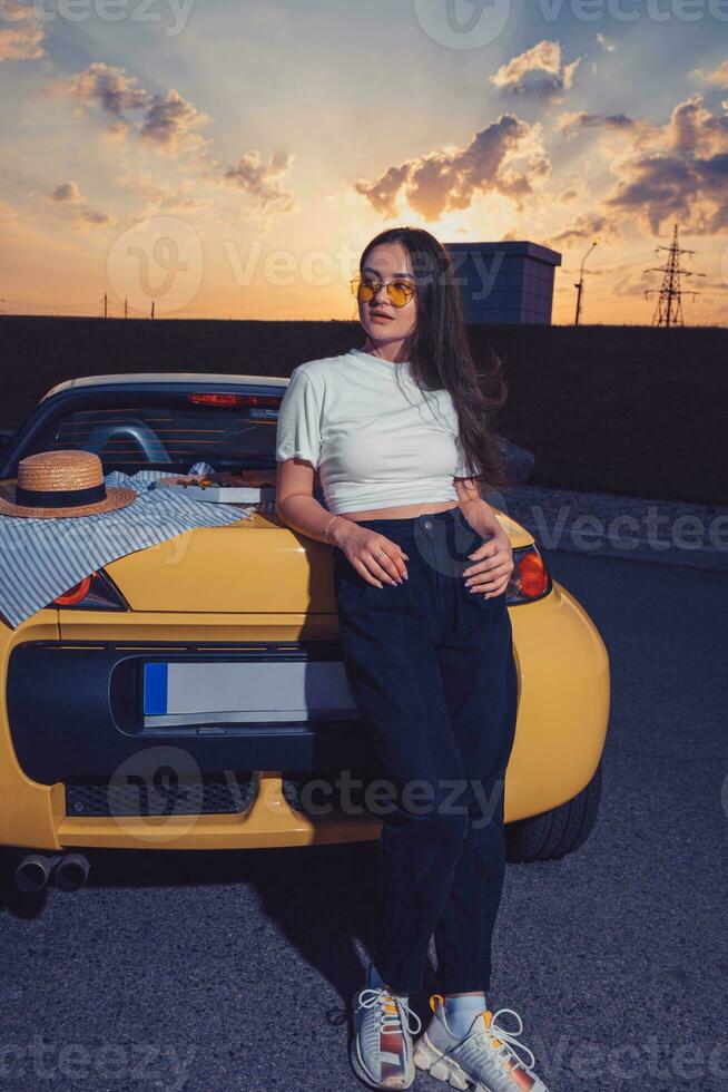 giovane signora nel occhiali da sole, bianca superiore, blu jeans e scarpe da ginnastica è in posa all'aperto vicino giallo auto roadster con Pizza su suo tronco. veloce cibo foto