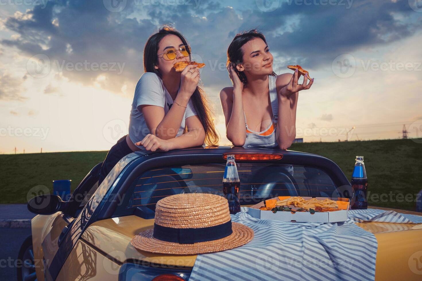 affascinante le signore nel casuale attrezzatura siamo mangiare Pizza, in posa nel giallo auto con francese patatine fritte, cappello e bibita nel bicchiere bottiglie su tronco. veloce cibo. finto su foto