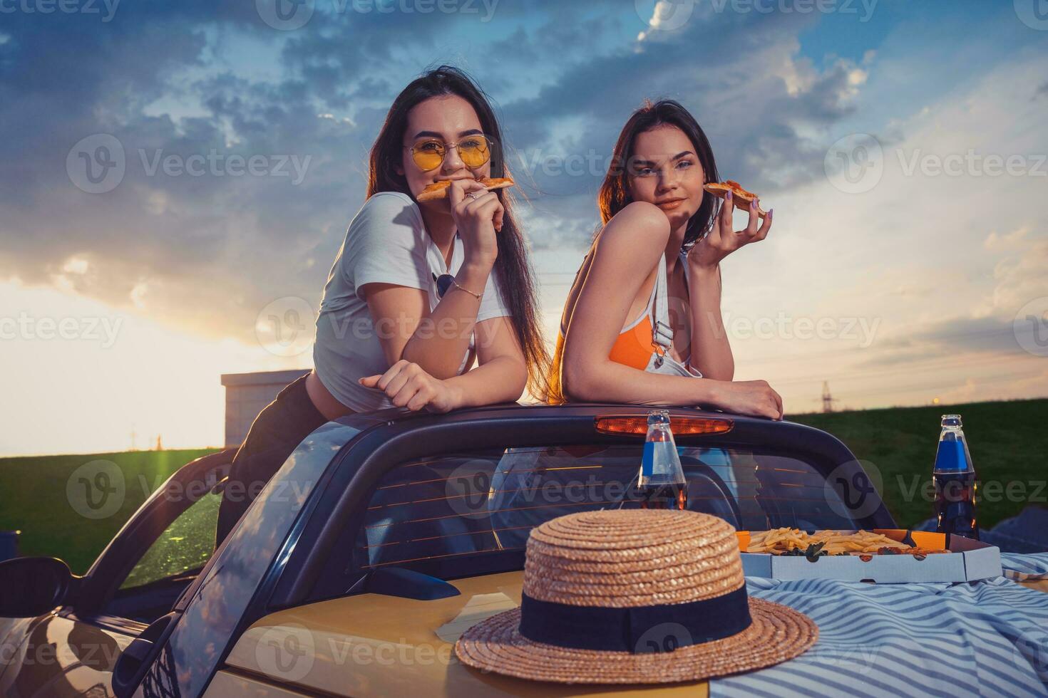 affascinante ragazze mangiare Pizza, in posa nel giallo auto roadster con francese patatine fritte, cappello e bibita nel bicchiere bottiglie su tronco. veloce cibo. finto su foto