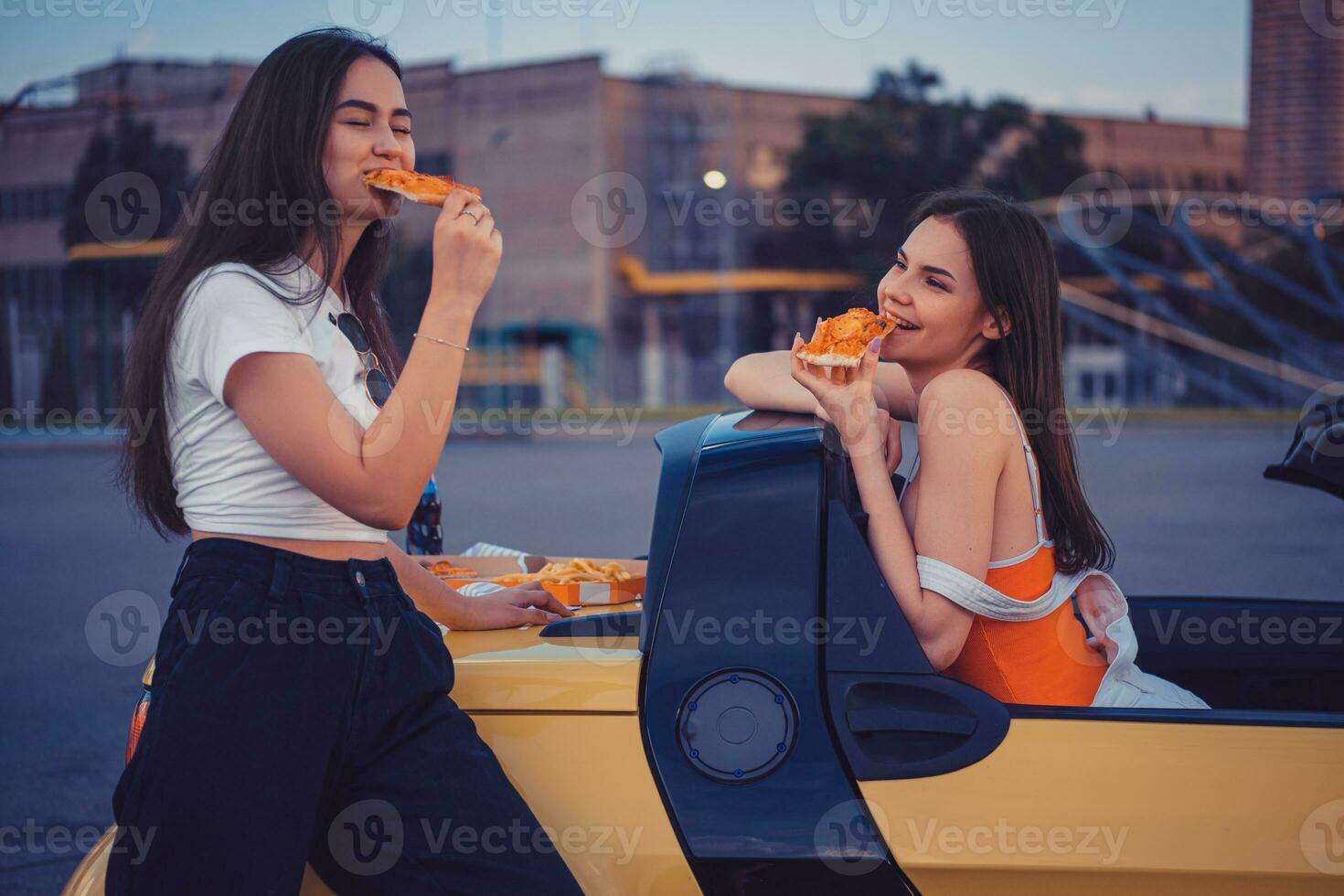 bella giovane donne nel casuale attrezzatura siamo mangiare Pizza mentre in posa nel giallo auto cabrio con francese patatine fritte e bibita acqua su suo tronco. veloce cibo foto